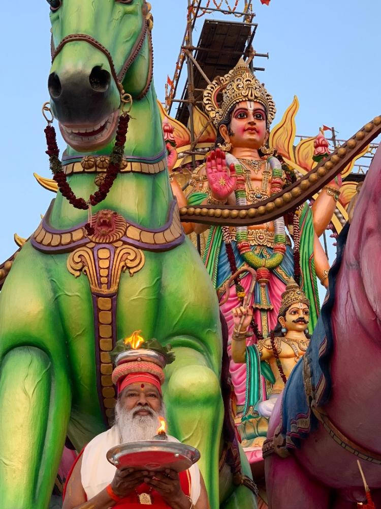 神 桌布 壁紙 — 一位祭司在節日期間，手持點亮的燈，立於一座騎著綠馬的宏偉彩色神像前。