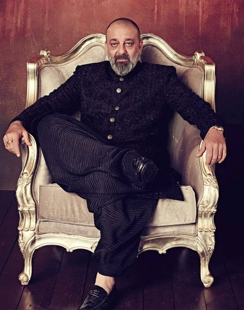 Sanjay 桌布 壁紙 — 一位禿頭灰鬍鬚的男人自信地坐在華麗的金色椅子上，背景是濃郁的紅牆。