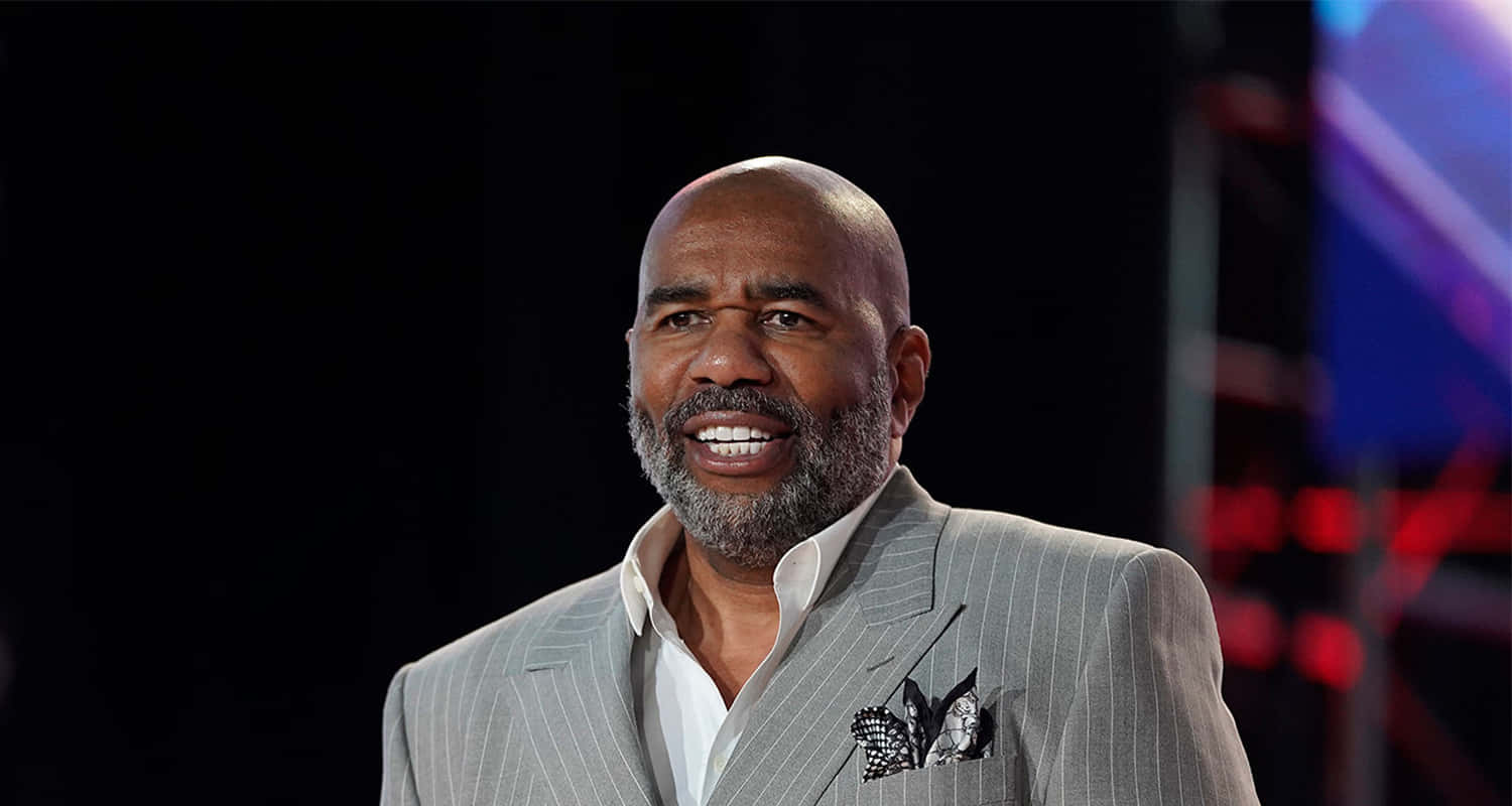 steve harvey 桌布 壁紙 — 一位禿頭灰鬍鬚的男子，身穿灰色細條紋西裝，在色彩繽紛燈光下的黑暗舞台上溫暖微笑。