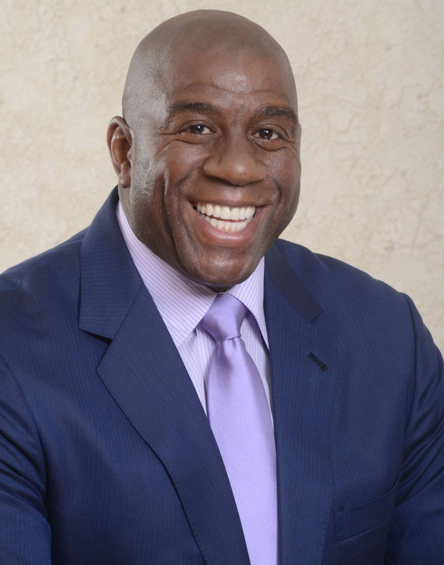 Magic Johnson 桌布 壁紙 — 一位禿頭男士穿著藍色西裝和薰衣草色領帶，對著有紋理的米色牆壁溫暖地微笑。