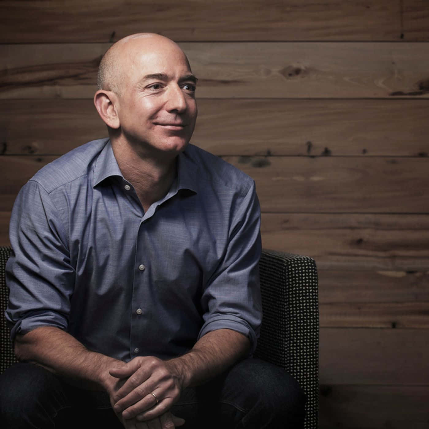 Jeff Bezos 桌布 壁紙 — 一位禿頭男子身穿藍色襯衫，坐在圖案椅子上，背景是溫暖的木牆，面帶溫柔的微笑。