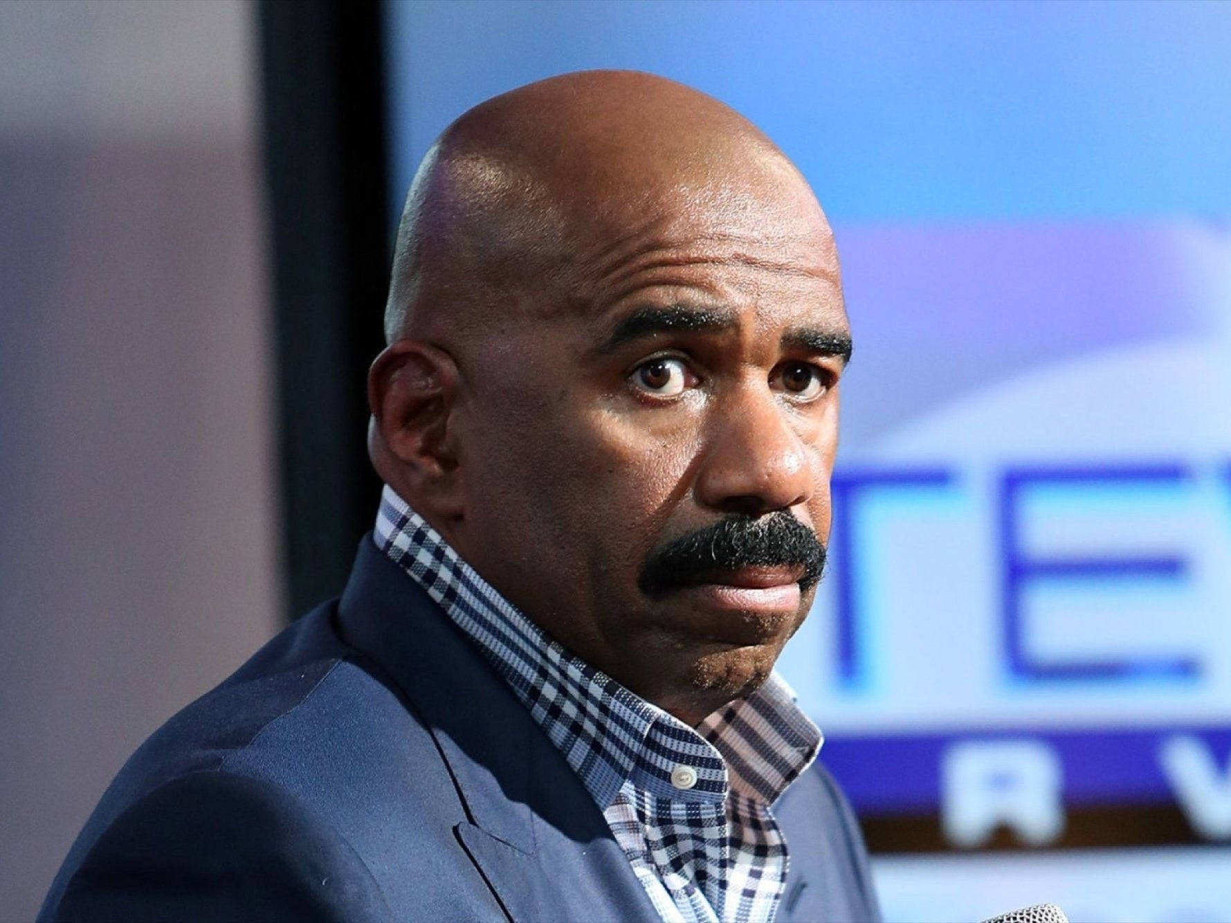 steve harvey 桌布 壁紙 — 一位禿頭留鬍鬚的男人穿著藍色西裝和格子襯衫，專注地向前看的特寫。