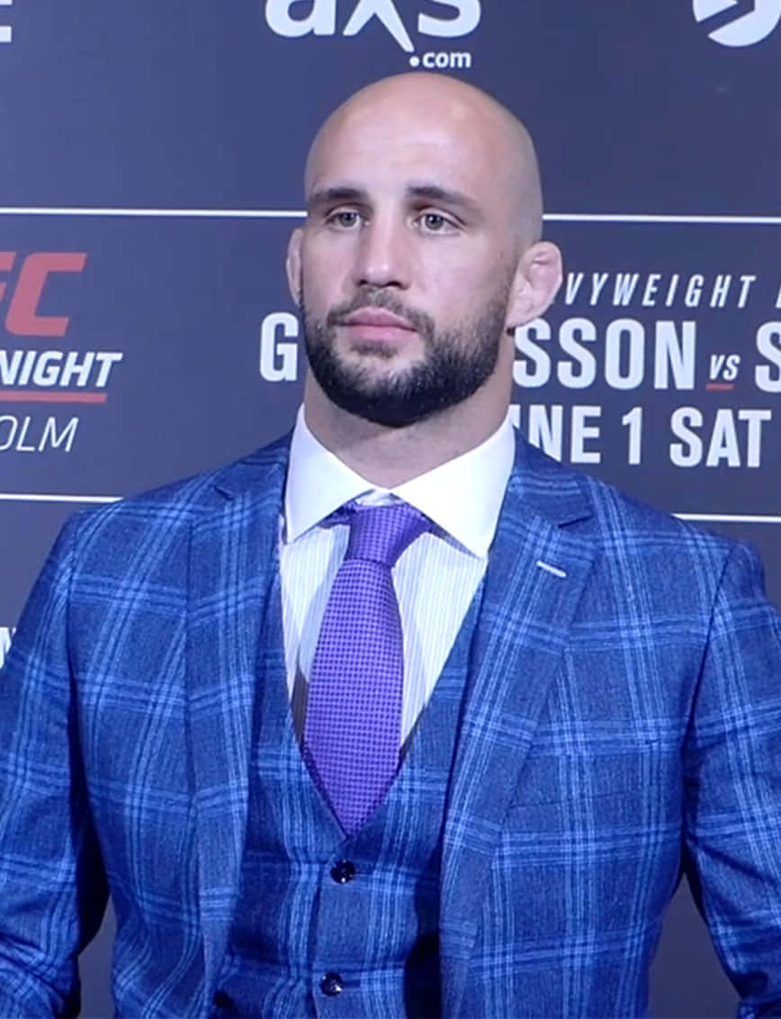MMA 桌布 壁紙 — 一位禿頭留鬍鬚的男子在UFC記者會上穿著藍色格紋西裝和紫色領帶。