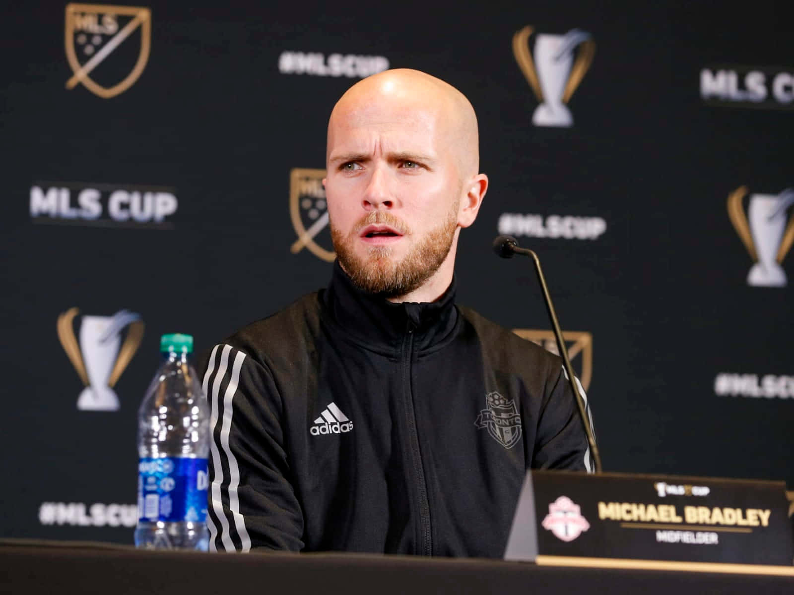 Athlete 桌布 壁紙 — 一位禿頭留鬍鬚的男子穿著黑色Adidas外套，身後有MLS Cup標誌，在新聞發布會上發言。