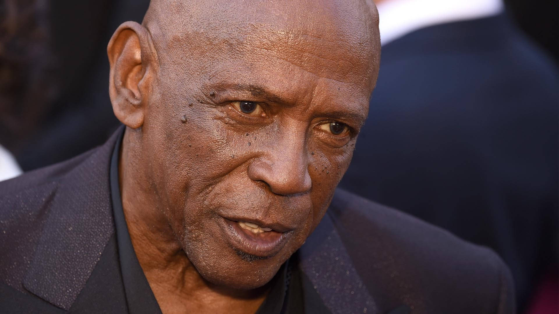Celebrity 桌布 壁紙 — 一位禿頭黑人男子，臉上帶有深深的皺紋，身穿深色西裝，稍微看向鏡頭外的特寫肖像。
