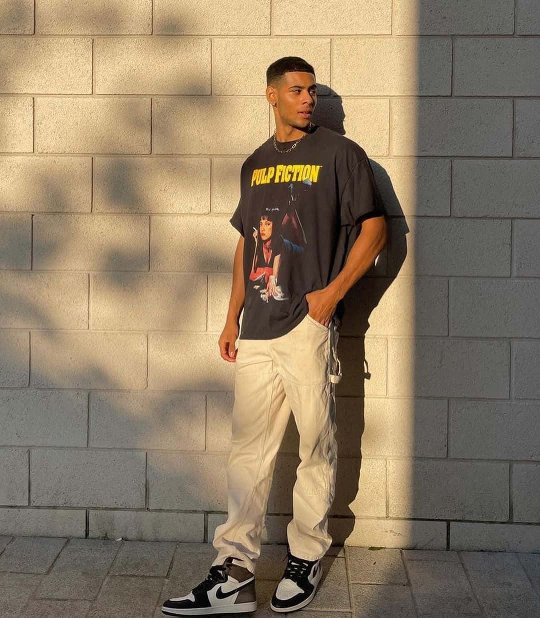 Black Clothing 桌布 壁紙 — 一位穿著 Pulp Fiction T恤和米色工裝褲的年輕男子，靠著陽光照射的混凝土牆，投下長長的影子。