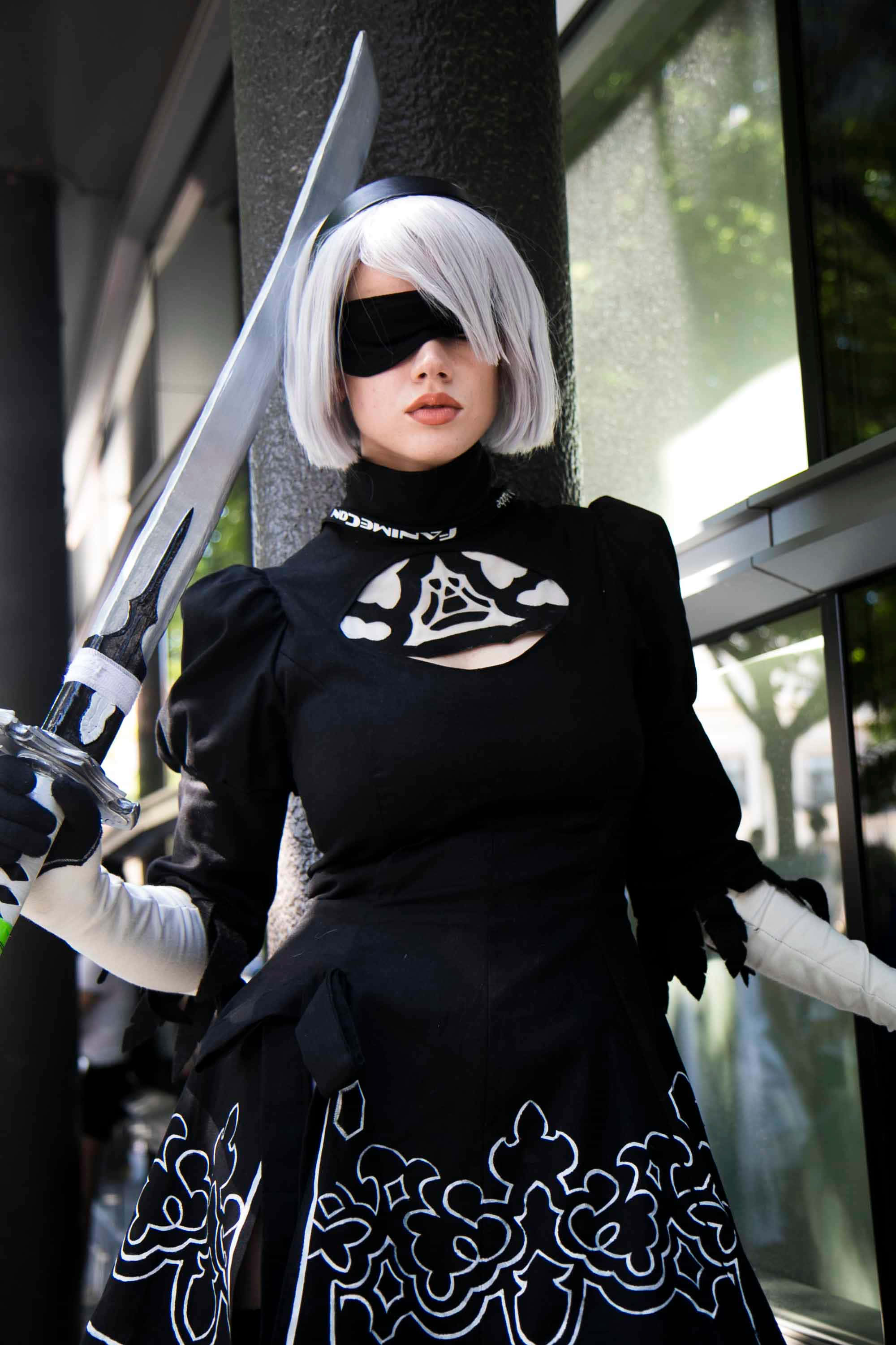 2B 桌布 壁紙 — 一位穿著《尼爾：自動人形》黑白2B服裝的角色扮演者，在現代建築背景下手持一把大劍。