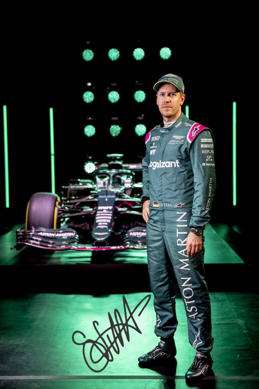 sebastian vettel 桌布 壁紙 — 一位穿著Aston Martin賽車裝備的F1車手，在閃爍的綠色舞台燈光下自信地站在車隊賽車前。