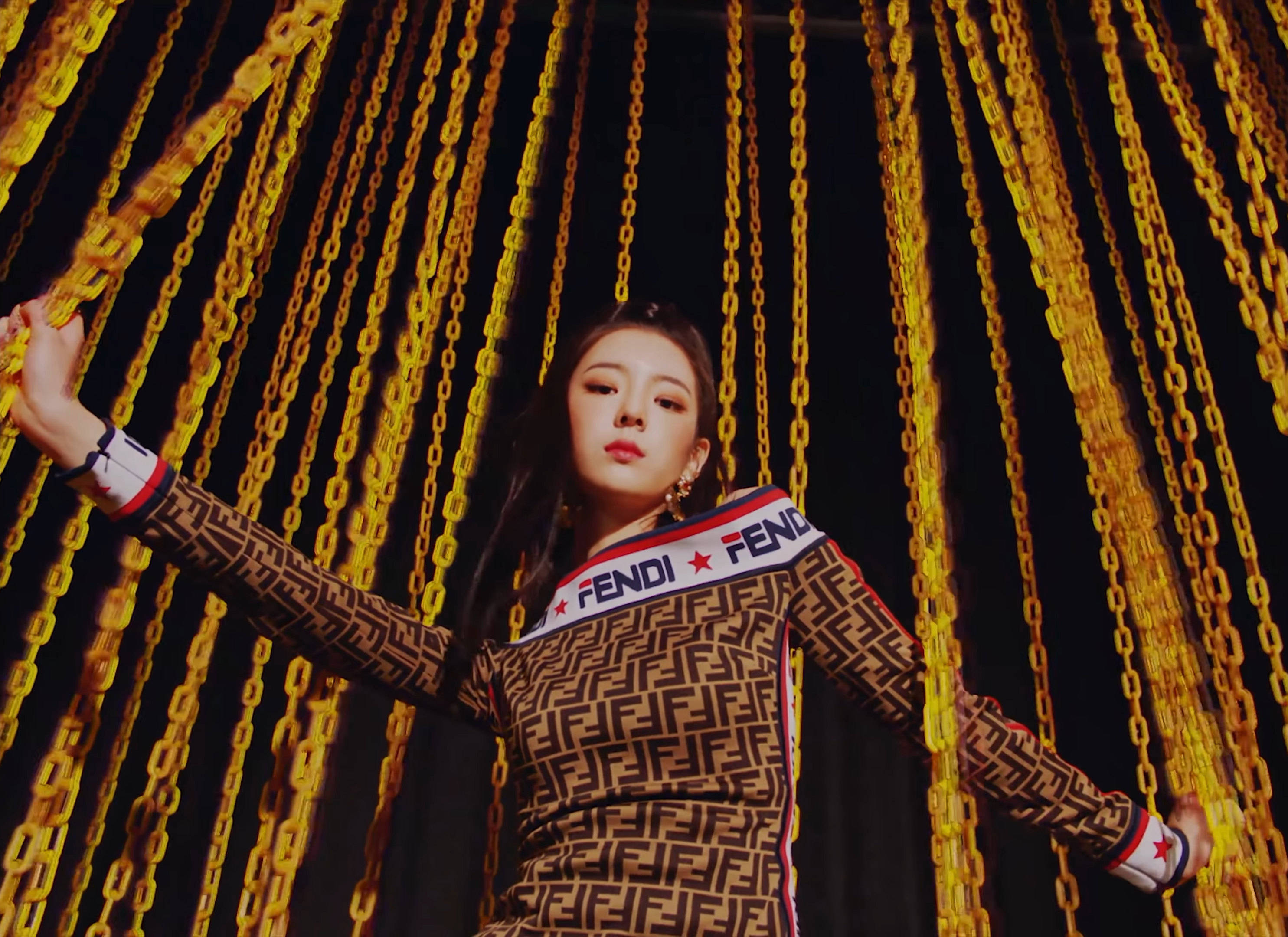 Lia 桌布 壁紙 — 一位穿著Fendi圖案服裝的女性，在黑暗背景下自信地站在層疊的金色鏈條之中。