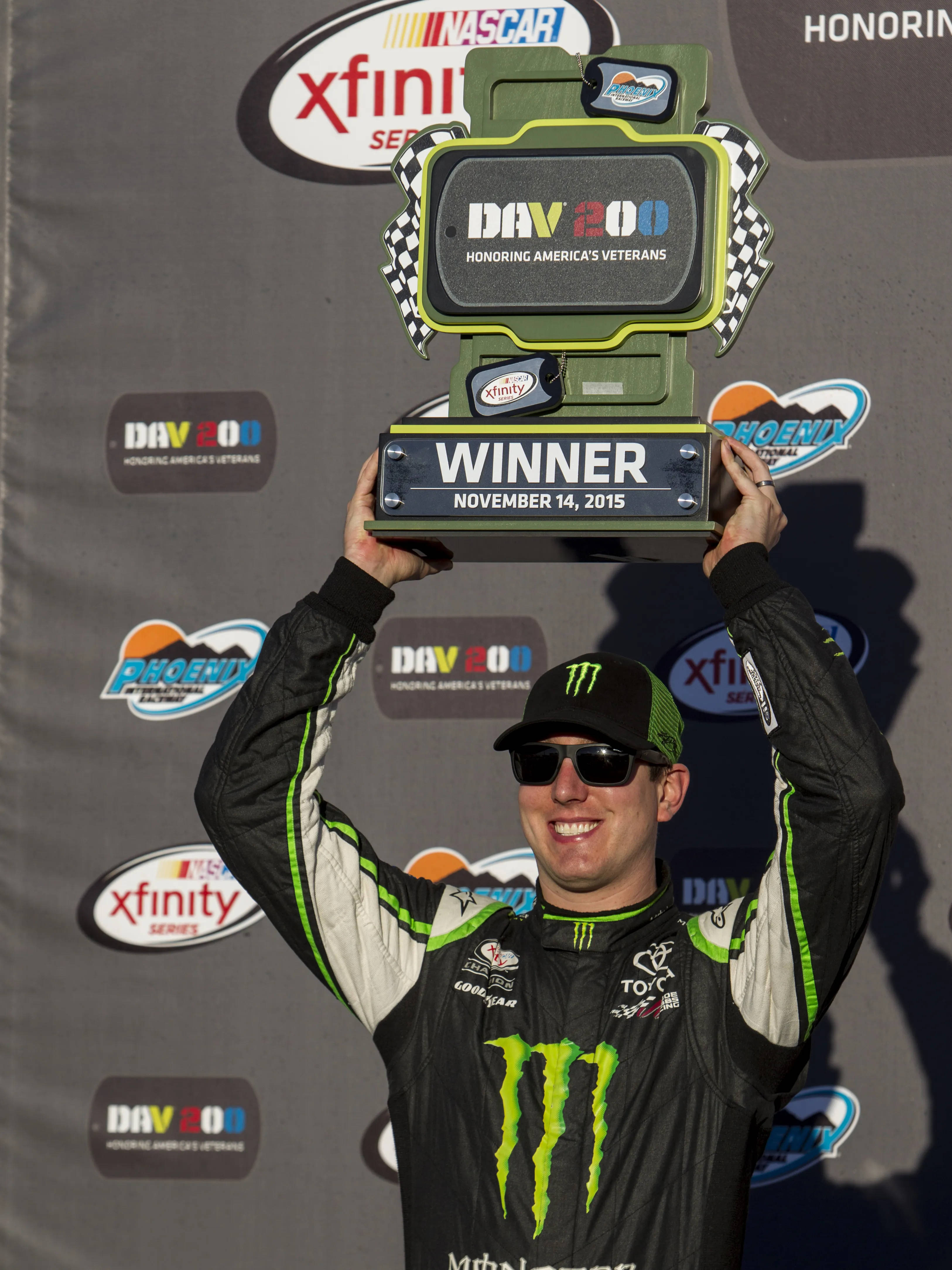 Trophy 桌布 壁紙 — 一位穿著Monster Energy制服的NASCAR車手在DAV 200比賽中，笑容燦爛地高舉一個大型綠色和黑色的「Winner」獎盃。