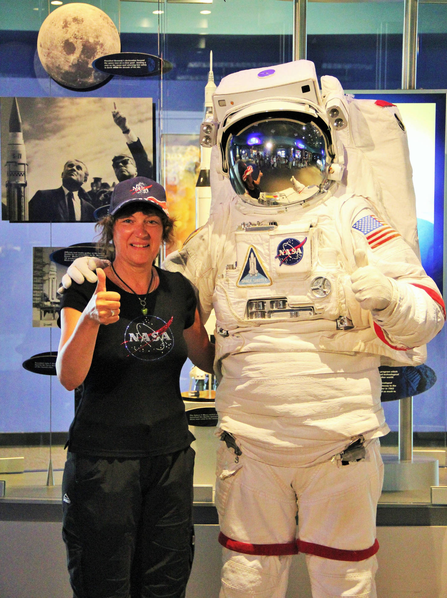  壁紙 — 一位穿著NASA T恤的微笑女士在博物館內一個真人大小的太空衣展品旁邊豎起大拇指。