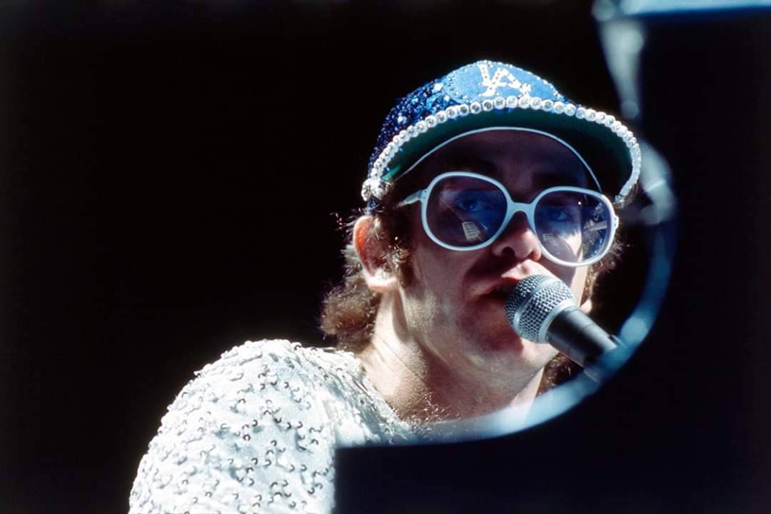 Rocketman 桌布 壁紙 — 一位穿著亮片服裝和超大太陽眼鏡的表演者在明亮的舞台燈光下對著麥克風唱歌。