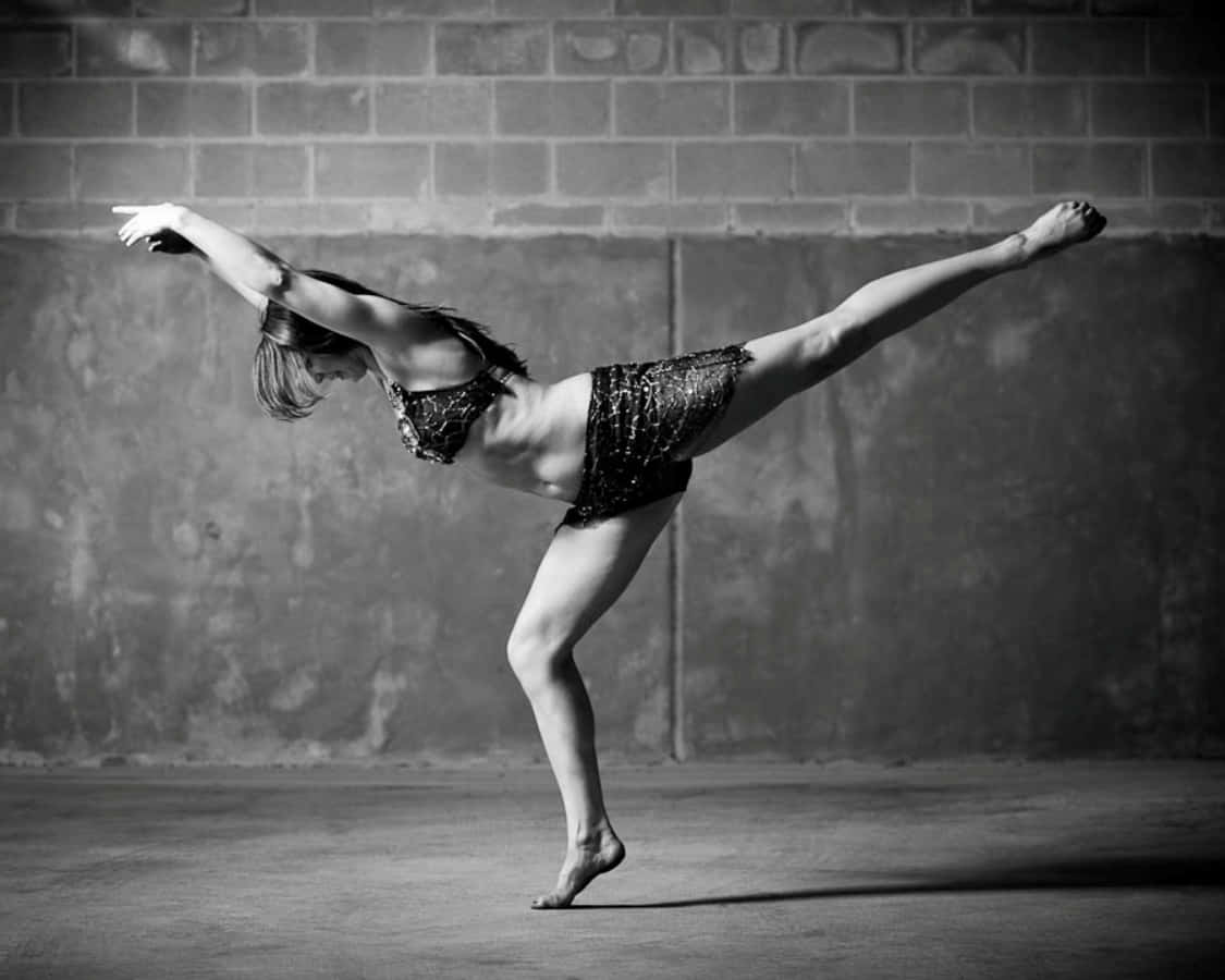 dancer 桌布 壁紙 — 一位穿著亮片服裝的舞者，在單色調的紋理磚牆前擺出優雅的阿拉貝斯克姿勢。