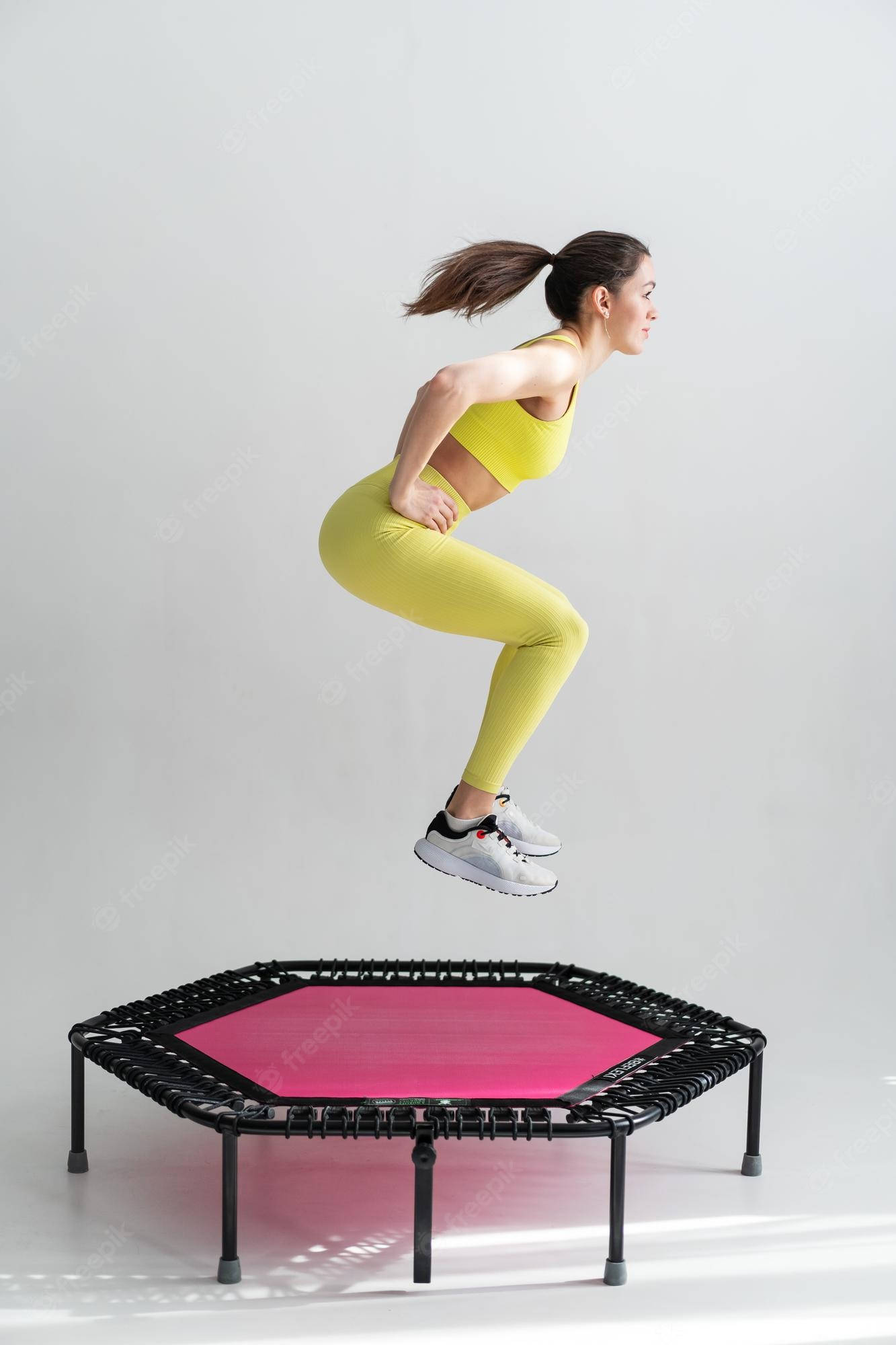 Exercise 桌布 壁紙 — 一位穿著亮黃色運動服的女士在白色純淨工作室背景前，於一個六角形粉紅色迷你彈跳床之上凌空跳躍。