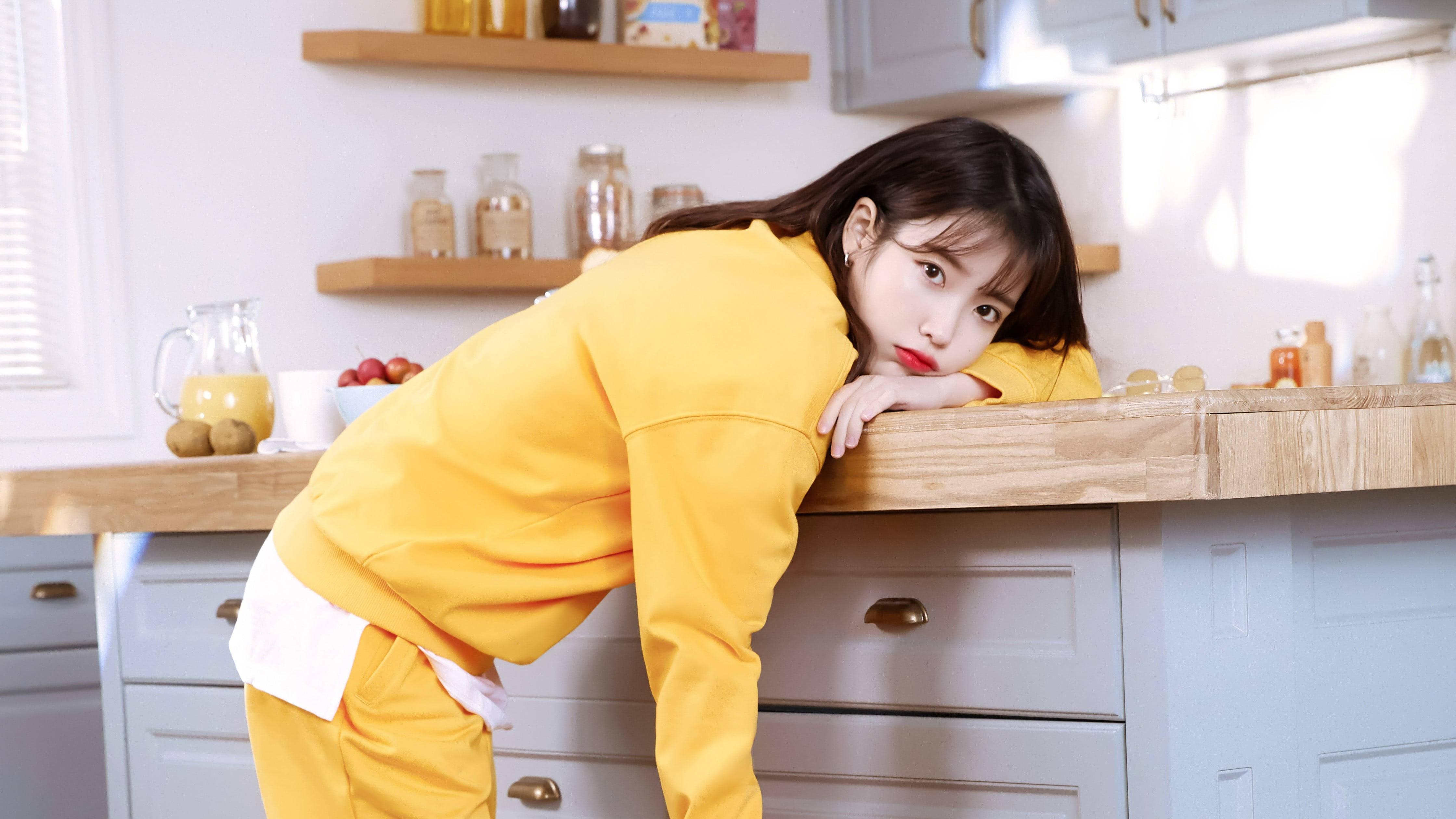 Cute Girl 桌布 壁紙 — 一位穿著亮黃色運動服的年輕女子，帶著俏皮的表情靠在木製廚房檯面上。