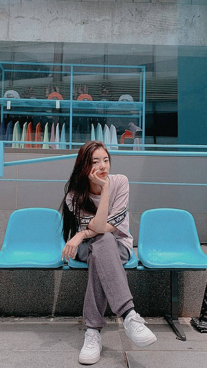 Lia 桌布 壁紙 — 一位穿著休閒街頭服的年輕女性，坐在明亮的藍色體育場座位上，背景是現代玻璃建築外牆，若有所思。