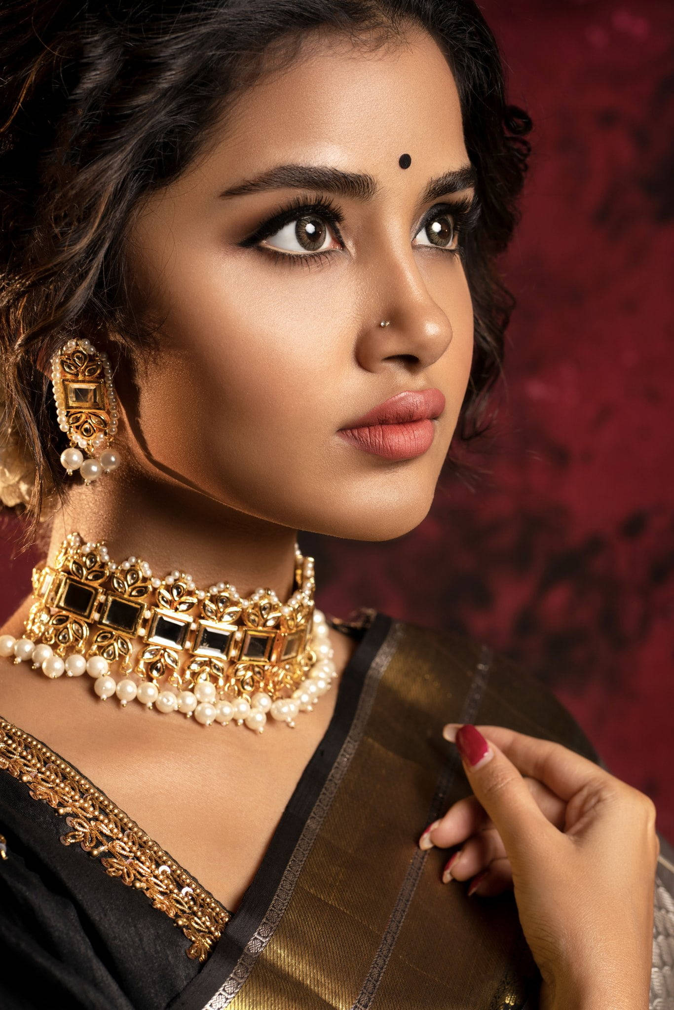 anupama parameswaran 桌布 壁紙 — 一位穿著傳統印度服飾、佩戴金和珍珠首飾的女性，在豐富的紅色紋理背景下若有所思地凝視。