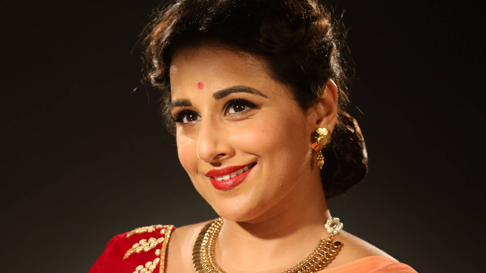 Vidya Balan 桌布 壁紙 — 一位穿著傳統印度服飾、光彩照人的女性在深色背景下溫暖地微笑。