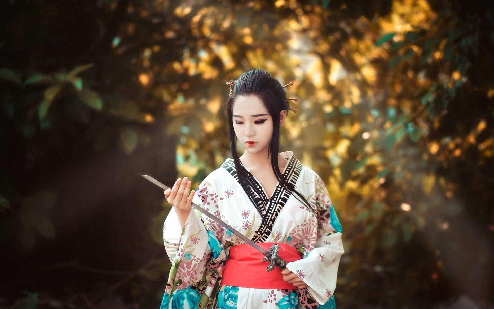 東京 桌布 壁紙 — 一位穿著傳統日本服飾的女性在陽光明媚、金色樹葉的森林中手持武士刀。