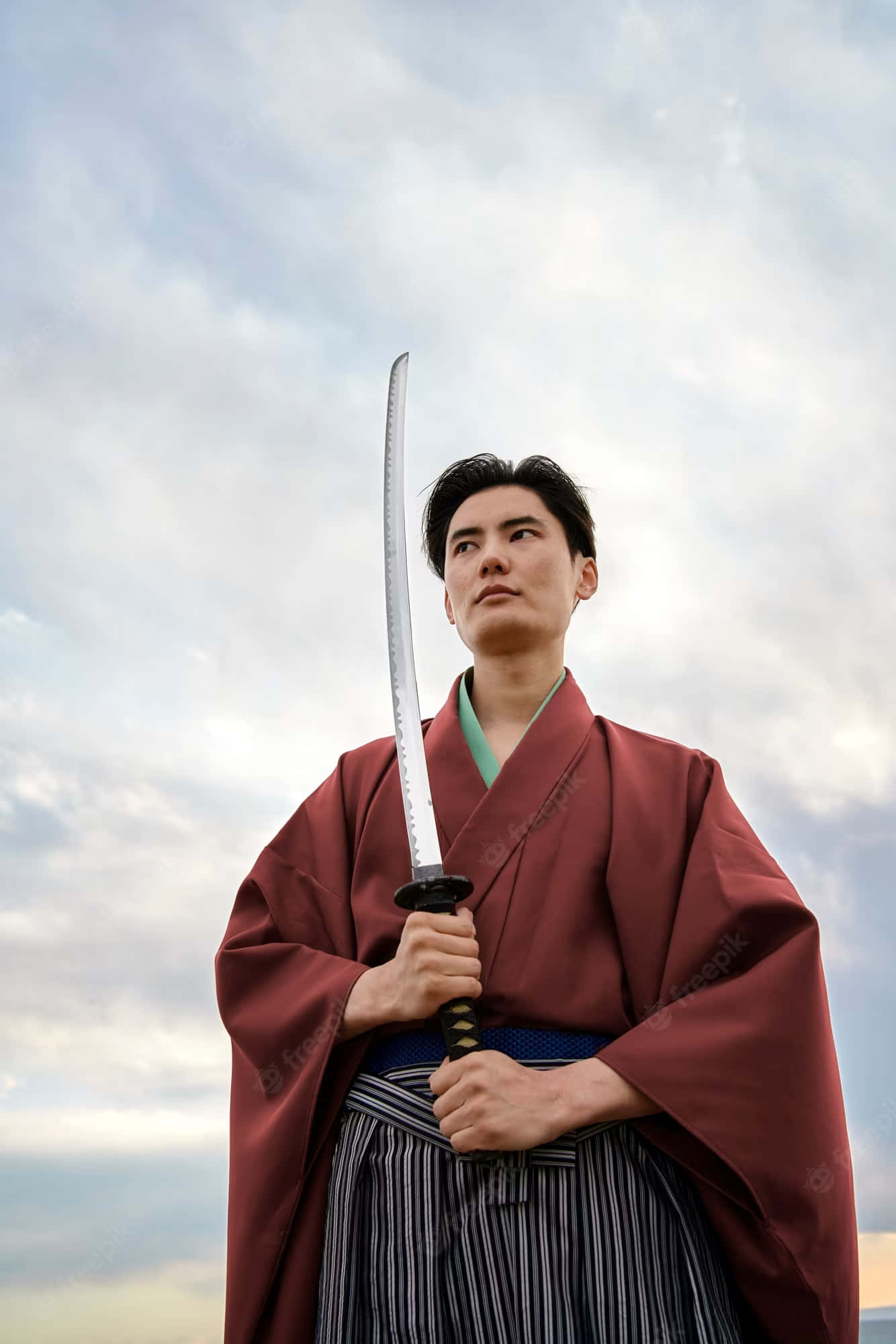 Samurai Sword 桌布 壁紙 — 一位穿著傳統日本服飾的年輕男子，在陰天的天空下，神情專注地握著一把武士刀。