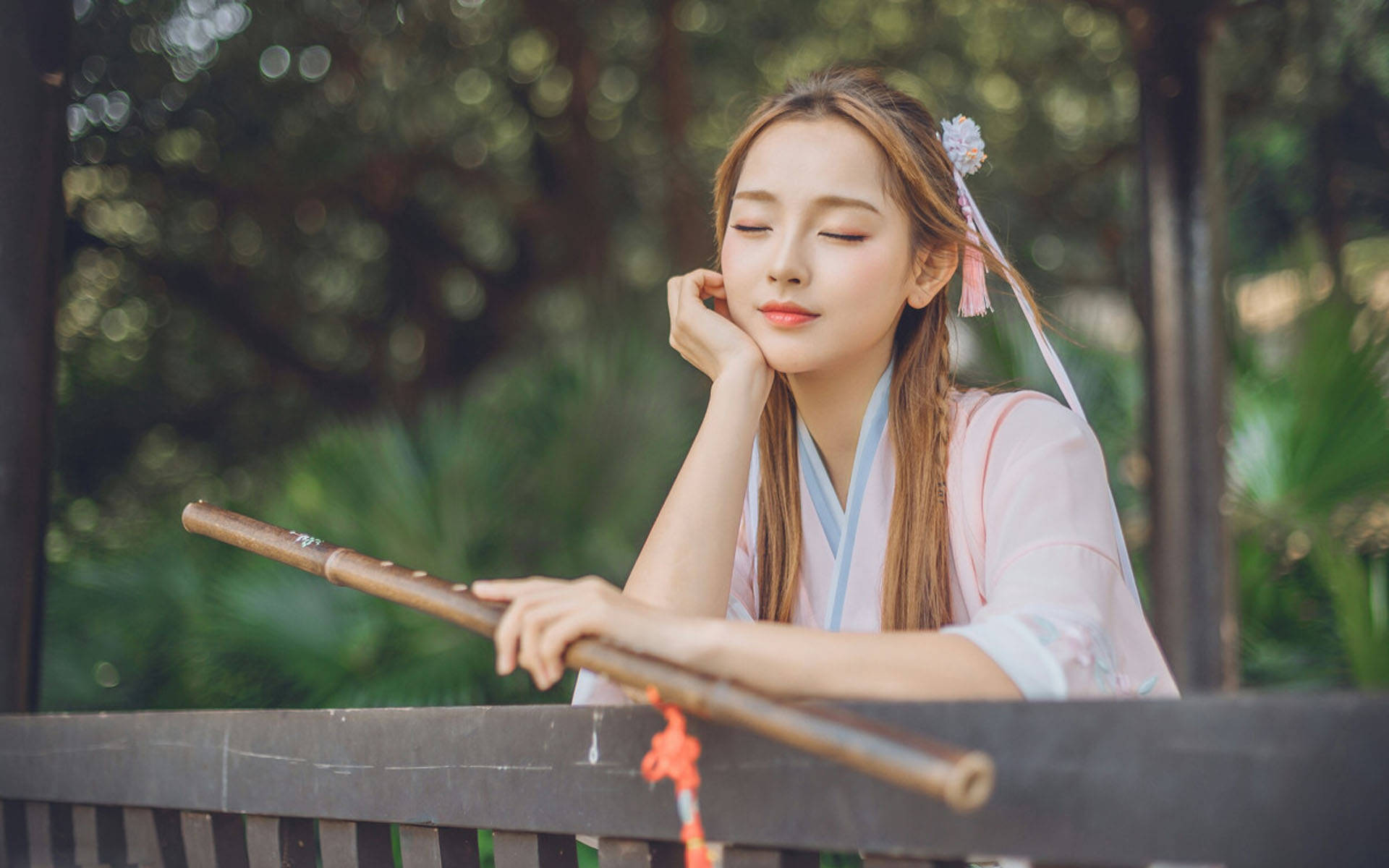 模特兒 桌布 壁紙 — 一位穿著傳統粉紅色服裝的寧靜女子，手托下巴，手持竹笛，背景是鬱鬱蔥蔥的花園。