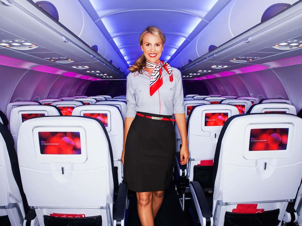 Flight Attendant 桌布 壁紙 — 一位穿著制服、面帶微笑的空中服務員在現代飛機艙的通道上行走，艙內有紫色環境燈光和紅色座椅背屏幕。