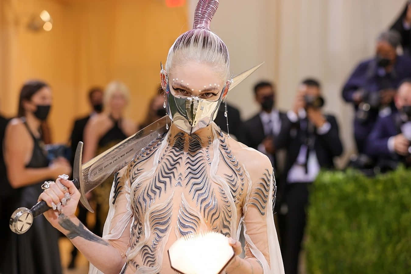 Met Gala 2021 桌布 壁紙 — 一位穿著前衛時尚、戴著金屬面具、手持劍的人士在華麗活動中擺姿勢。