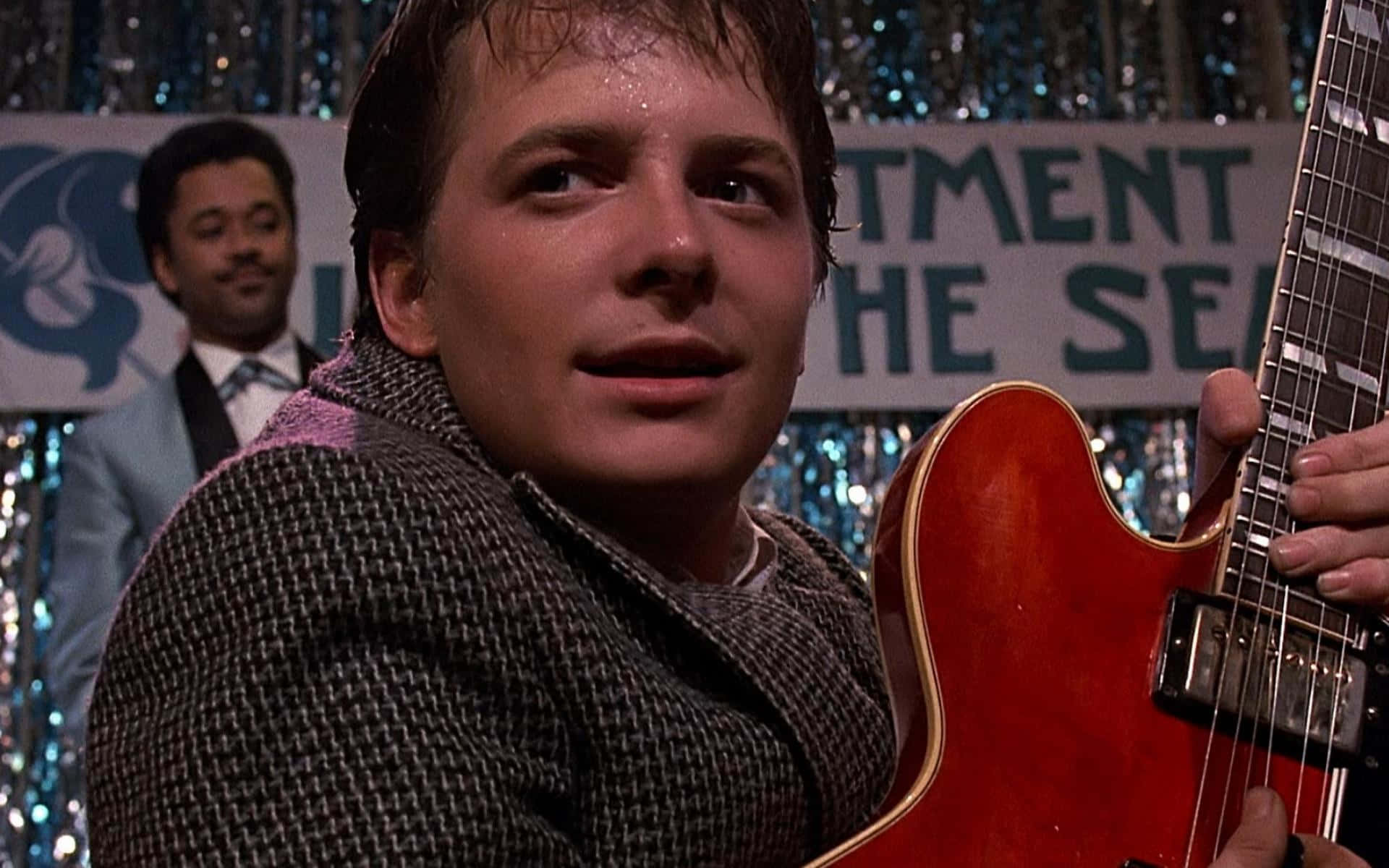 michael j fox 桌布 壁紙 — 一位穿著千鳥格外套的年輕男子，在閃爍的背景前拿著一把紅色電吉他，背景有一條寫著「TMENT HE SE」的橫幅，他身後還有另一位穿西裝的男子。