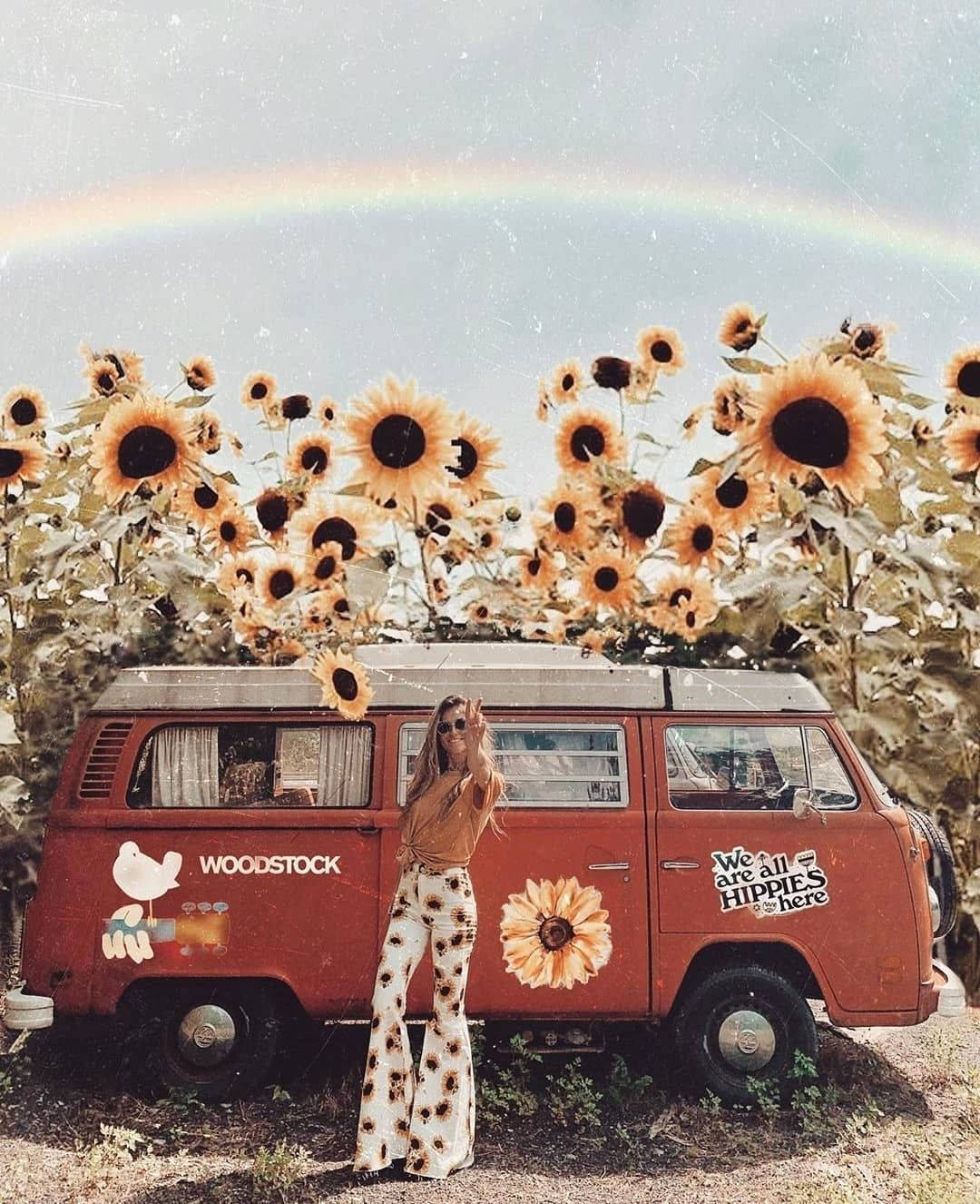 Woodstock 桌布 壁紙 — 一位穿著向日葵印花褲子的女士在一輛裝飾著嬉皮貼紙的復古紅色VW巴士旁揮手，彩虹在向日葵田上空拱起。