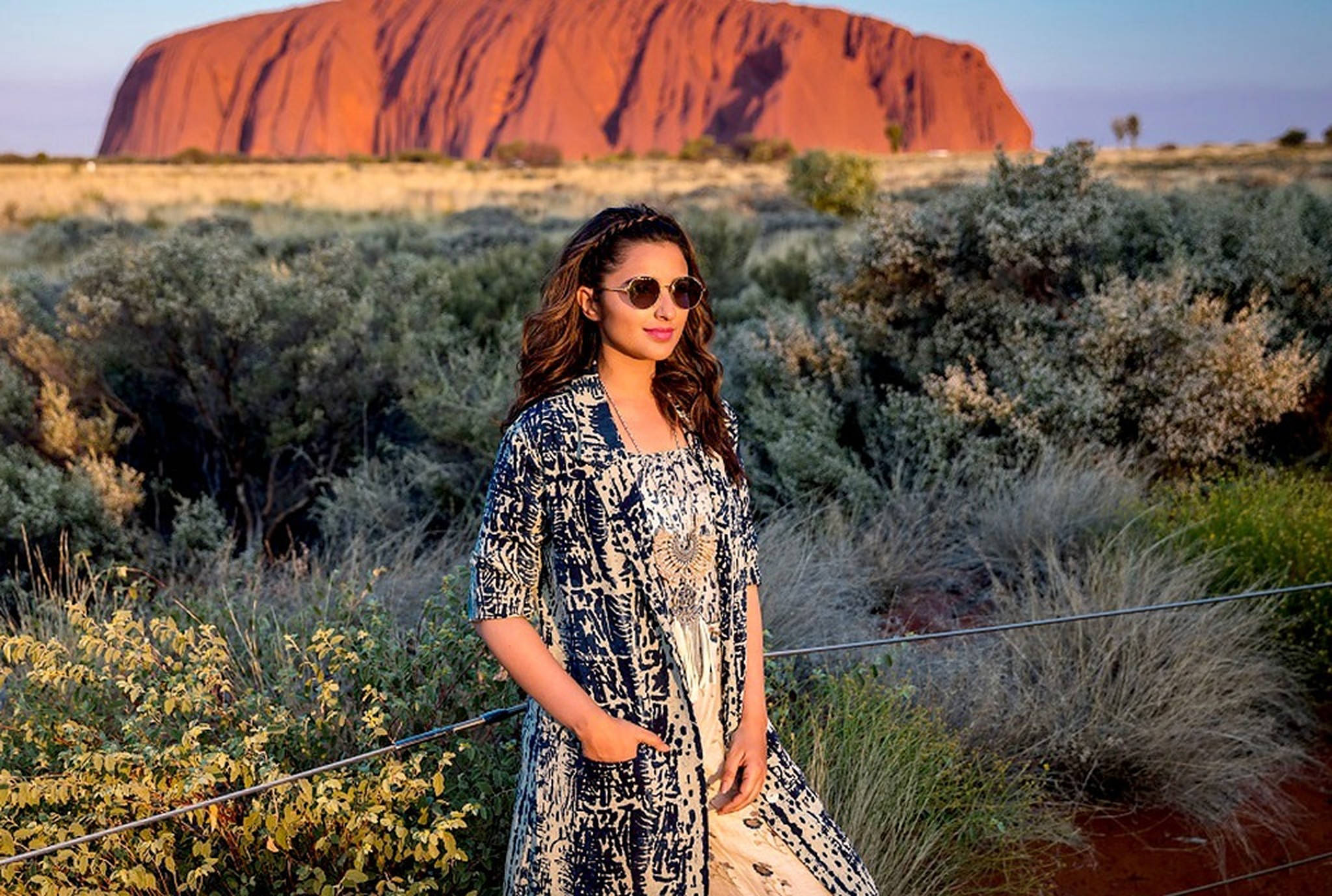 Parineeti 桌布 壁紙 — 一位穿著圖案服裝的女性在黃金時段站在烏魯魯前，背景有沙漠灌木和溫暖的紅色岩石。