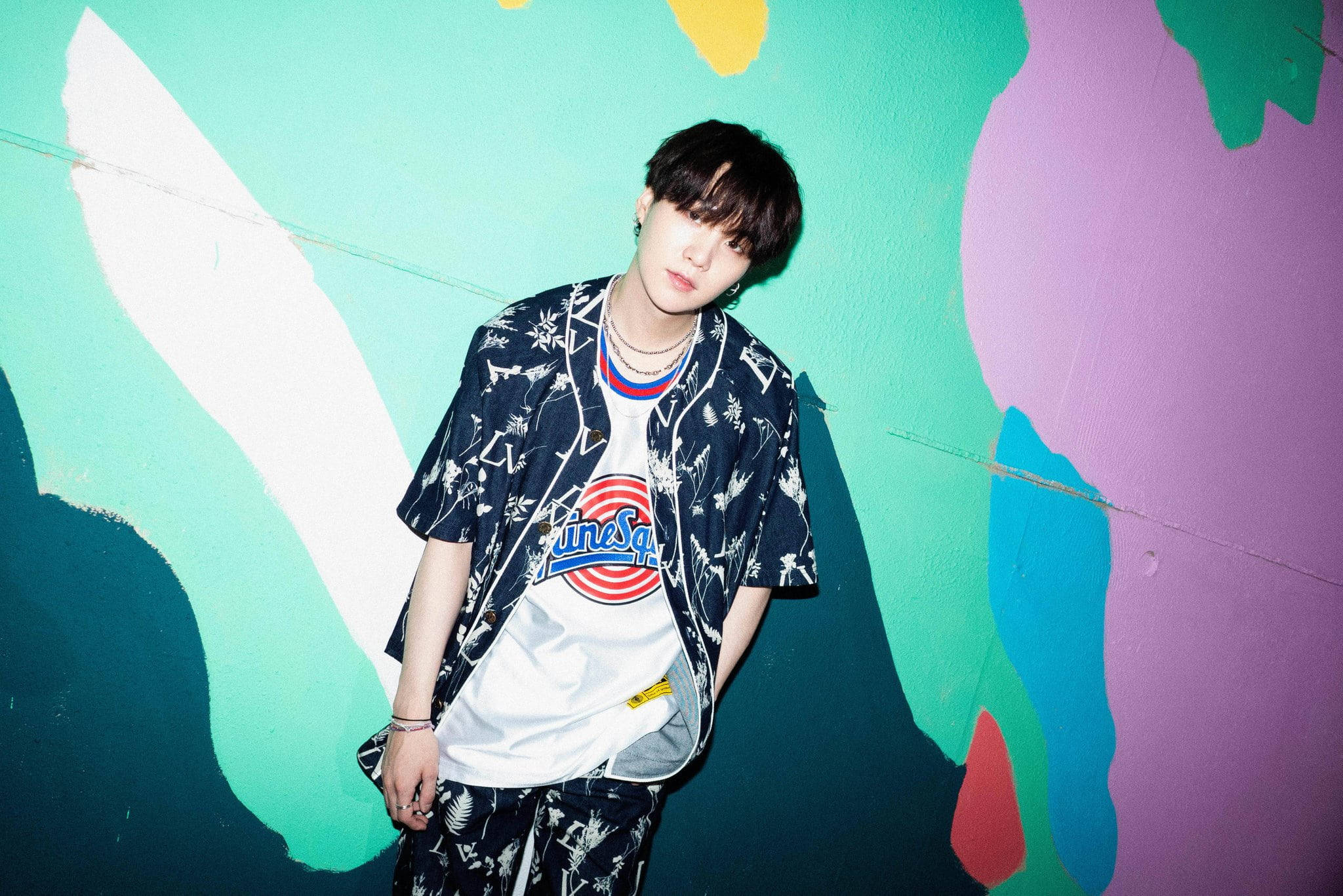 Bts Suga Aesthetic 桌布 壁紙 — 一位穿著圖案服裝的年輕男子自信地站在充滿活力的抽象壁畫牆前。