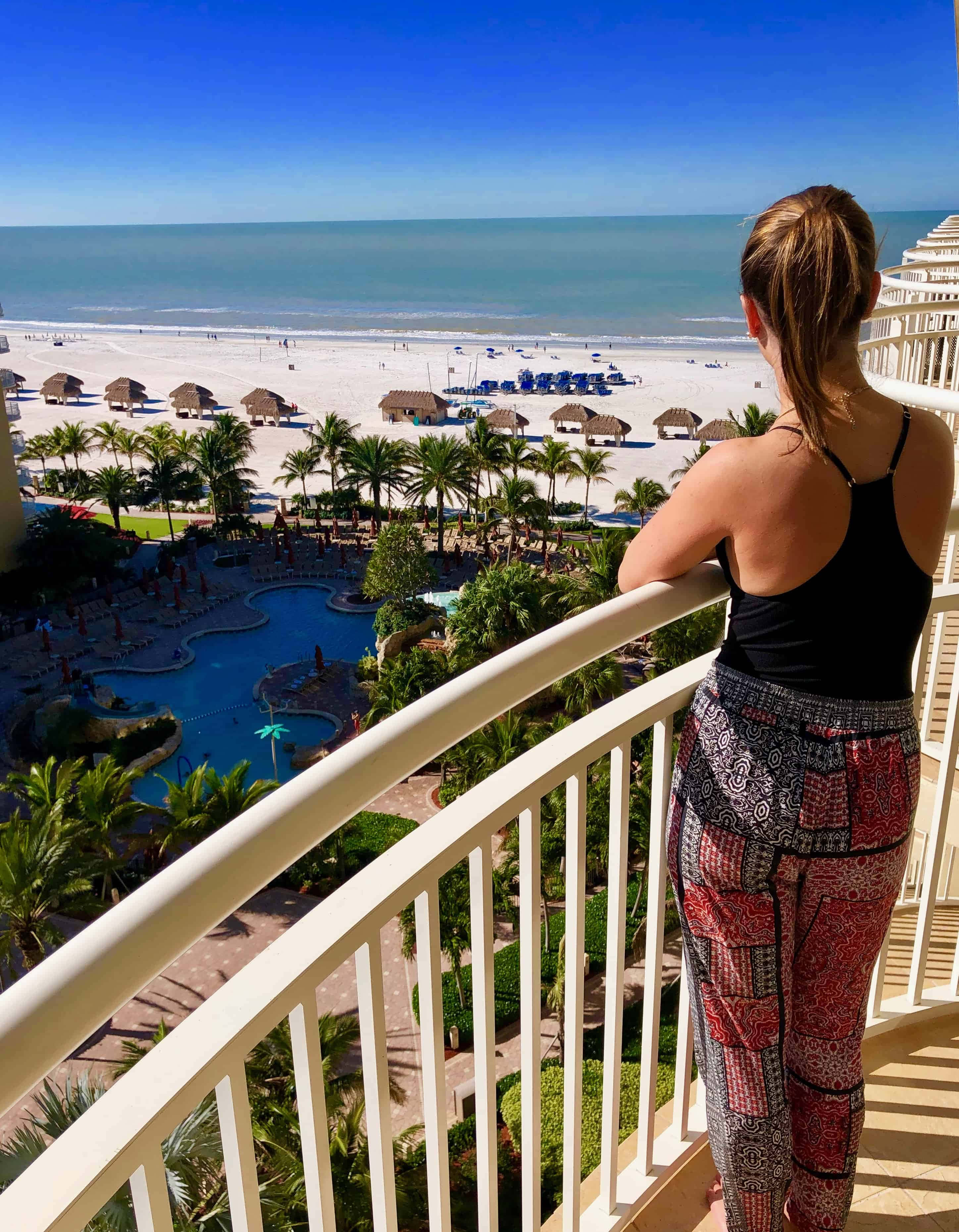 Marco Island 桌布 壁紙 — 一位穿著圖案褲子的女士站在陽台上，俯瞰著一個充滿活力的海灘度假村，那裡有碧藍的游泳池、棕櫚樹和寬闊的沙灘，天空清澈湛藍。