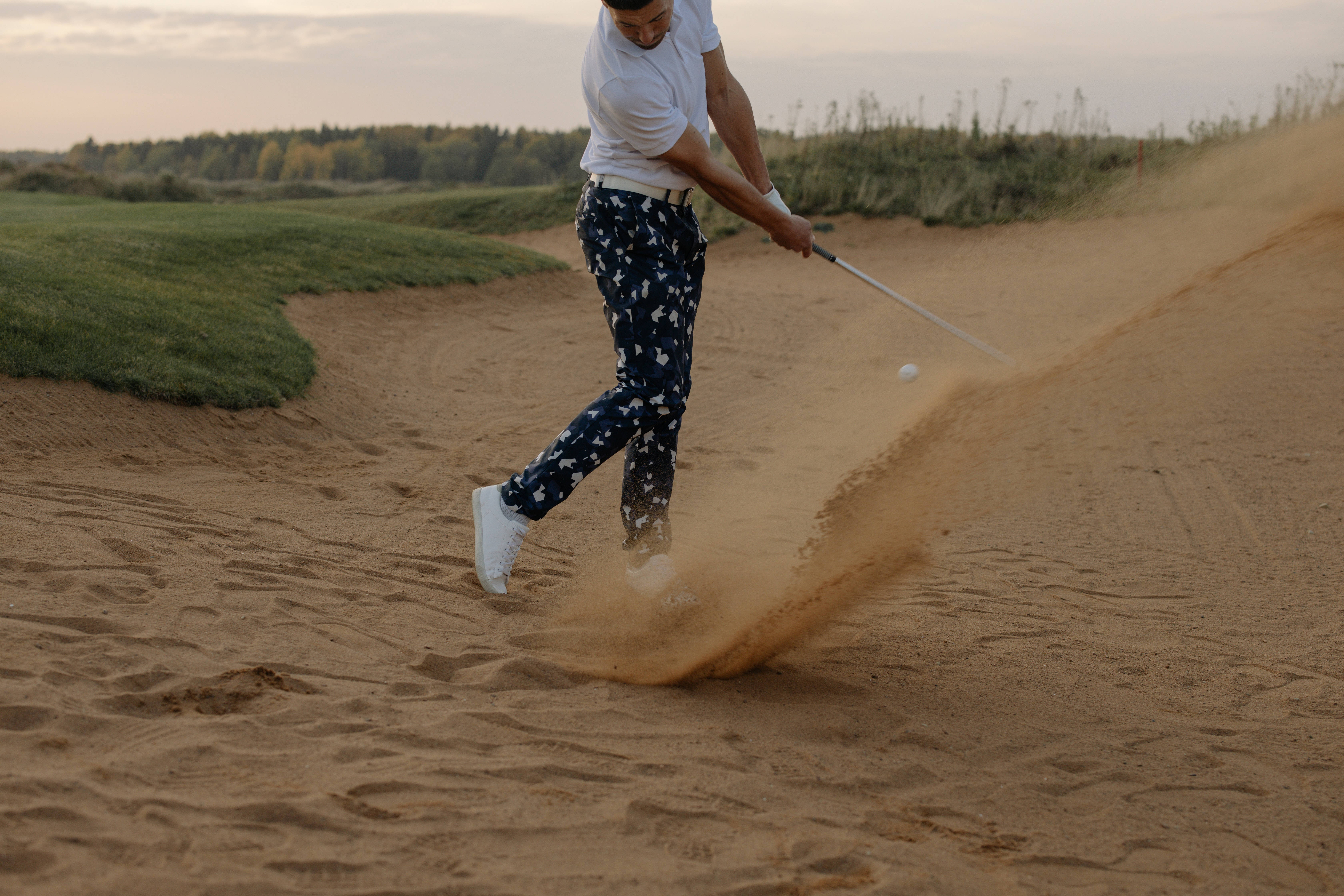 4K Golf 桌布 壁紙 — 一位穿著圖案褲子的高爾夫球手在日落時分揮桿穿過沙坑，揚起沙塵。