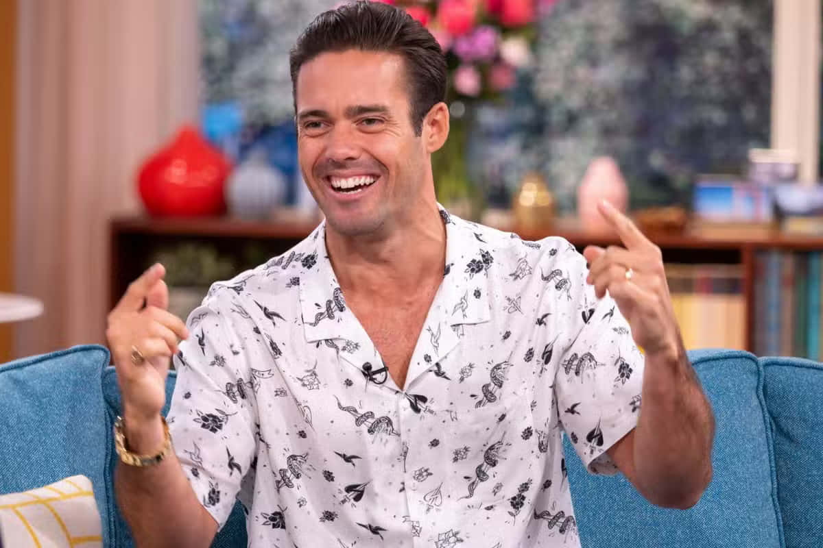 Spencer Matthews 桌布 壁紙 — 一位穿著圖案襯衫的開朗男子在明亮的攝影棚中，坐在藍色沙發上笑著做手勢。