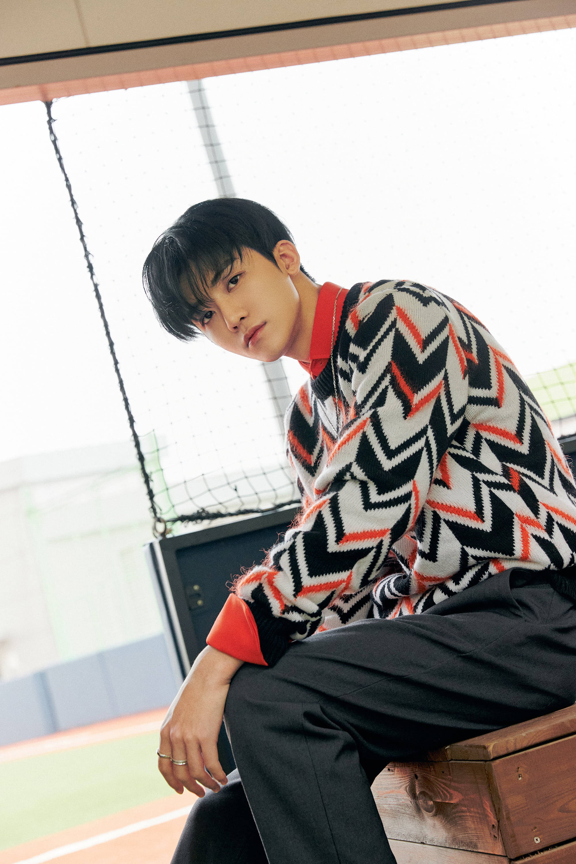 jaemin 桌布 壁紙 — 一位穿著大膽鋸齒紋毛衣的年輕男子，若有所思地坐在運動網附近的木長椅上。
