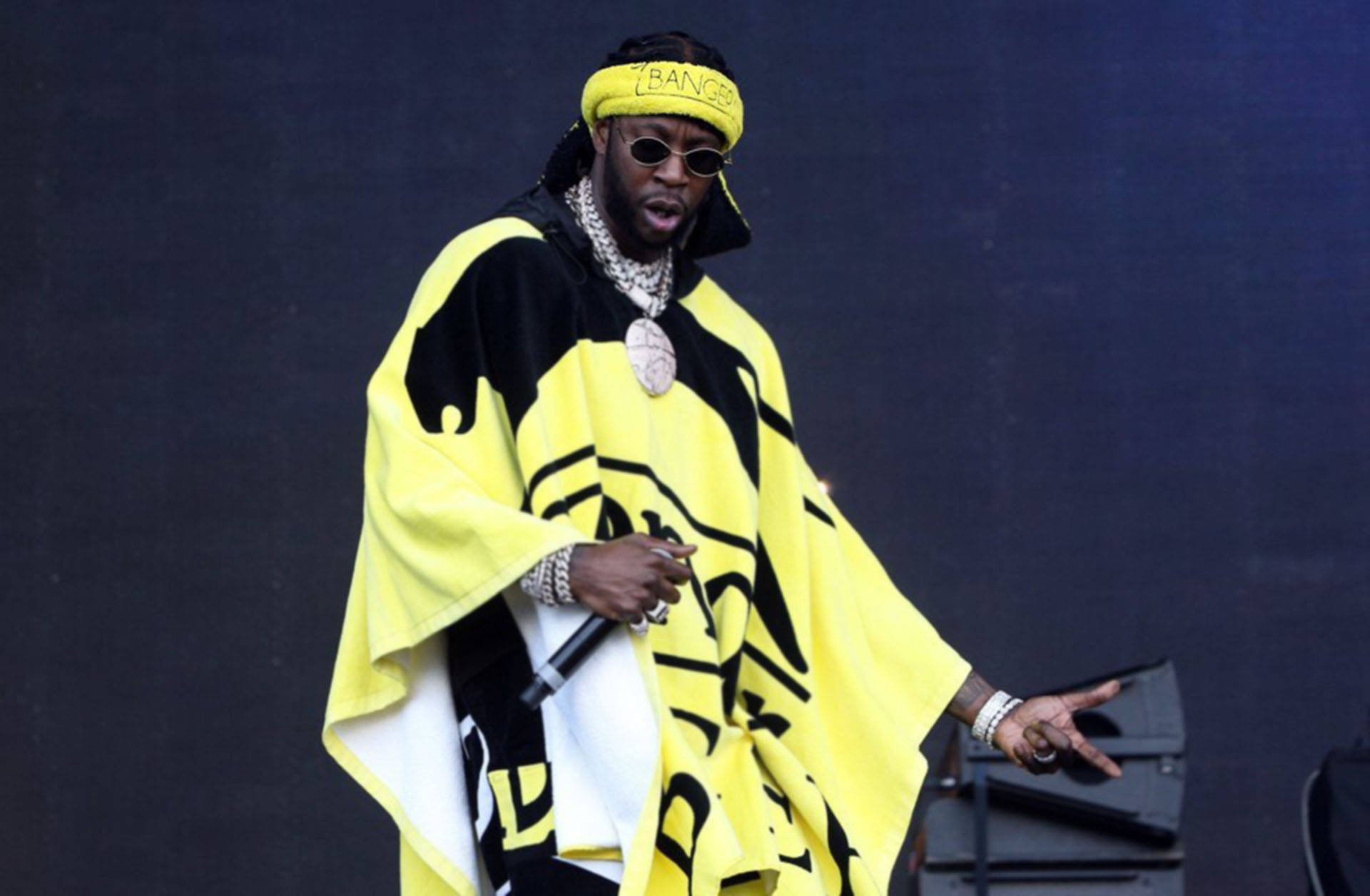 2 Chainz 桌布 壁紙 — 一位穿著大膽黃黑圖案斗篷的饒舌歌手，帶著麥克風和太陽眼鏡在舞台上表演。