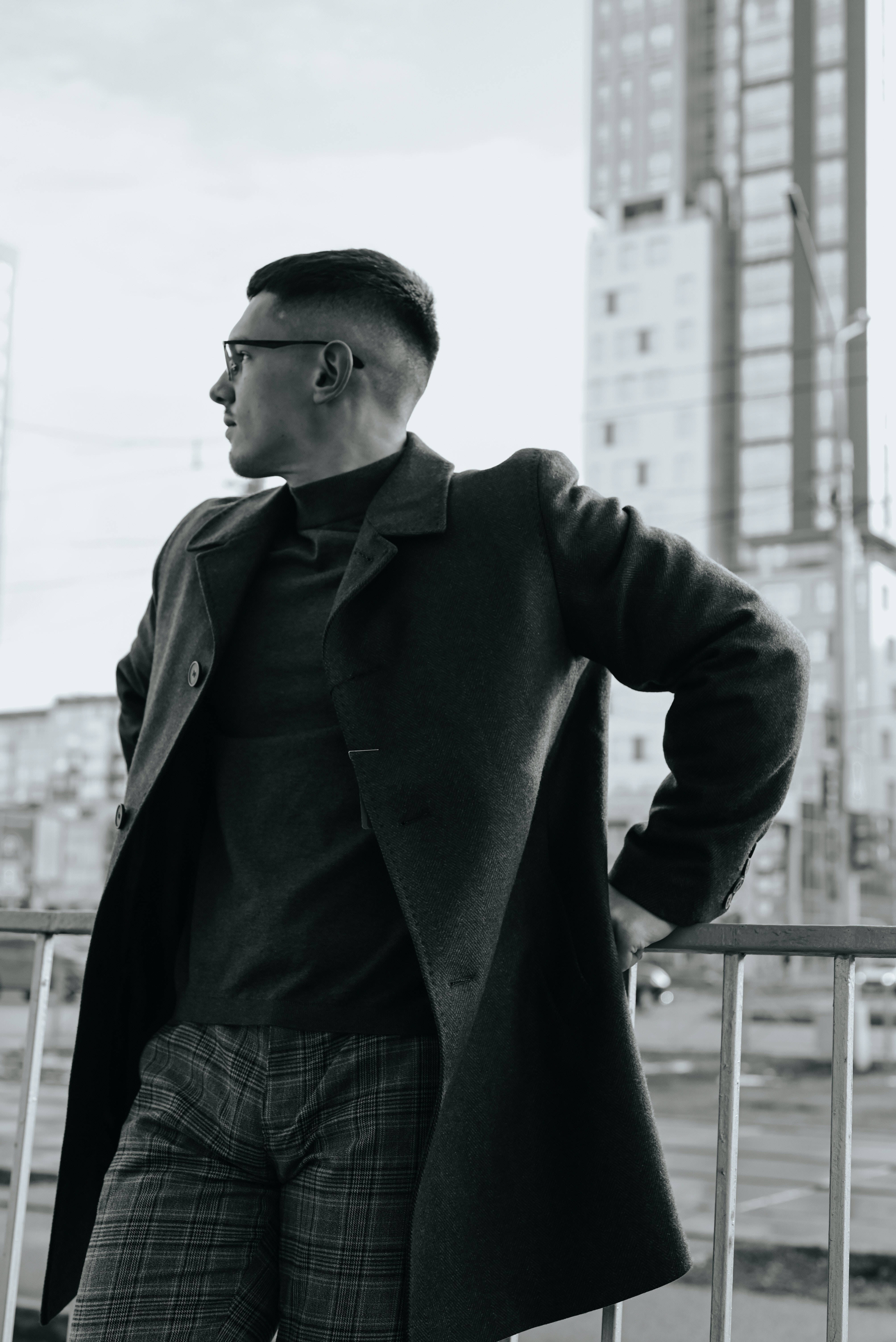 Man Aesthetic 桌布 壁紙 — 一位穿著大衣和眼鏡的時尚男士自信地站在城市陽台上，俯瞰單色調的高樓大廈。