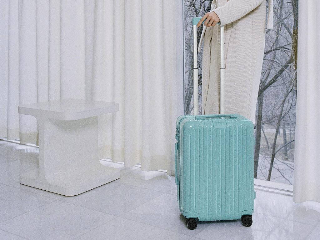 時尚 桌布 壁紙 — 一位穿著奶油色外套的人，在窗外有雪樹的窗邊拉著一個薄荷綠色的行李箱。