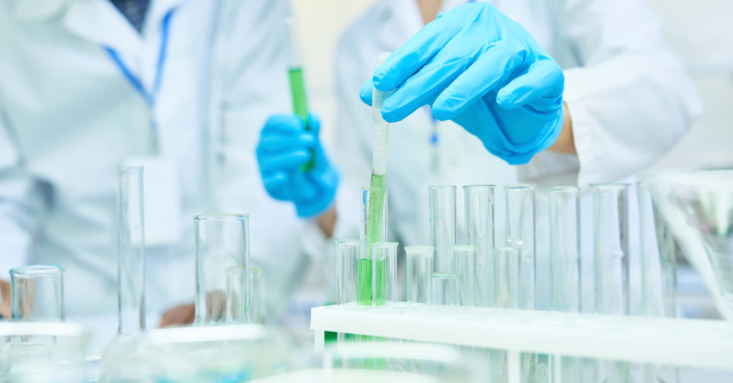 科學 桌布 壁紙 — 一位穿著實驗室外套和藍色手套的科學家，在其他玻璃器皿中小心處理一支裝有綠色液體的試管。