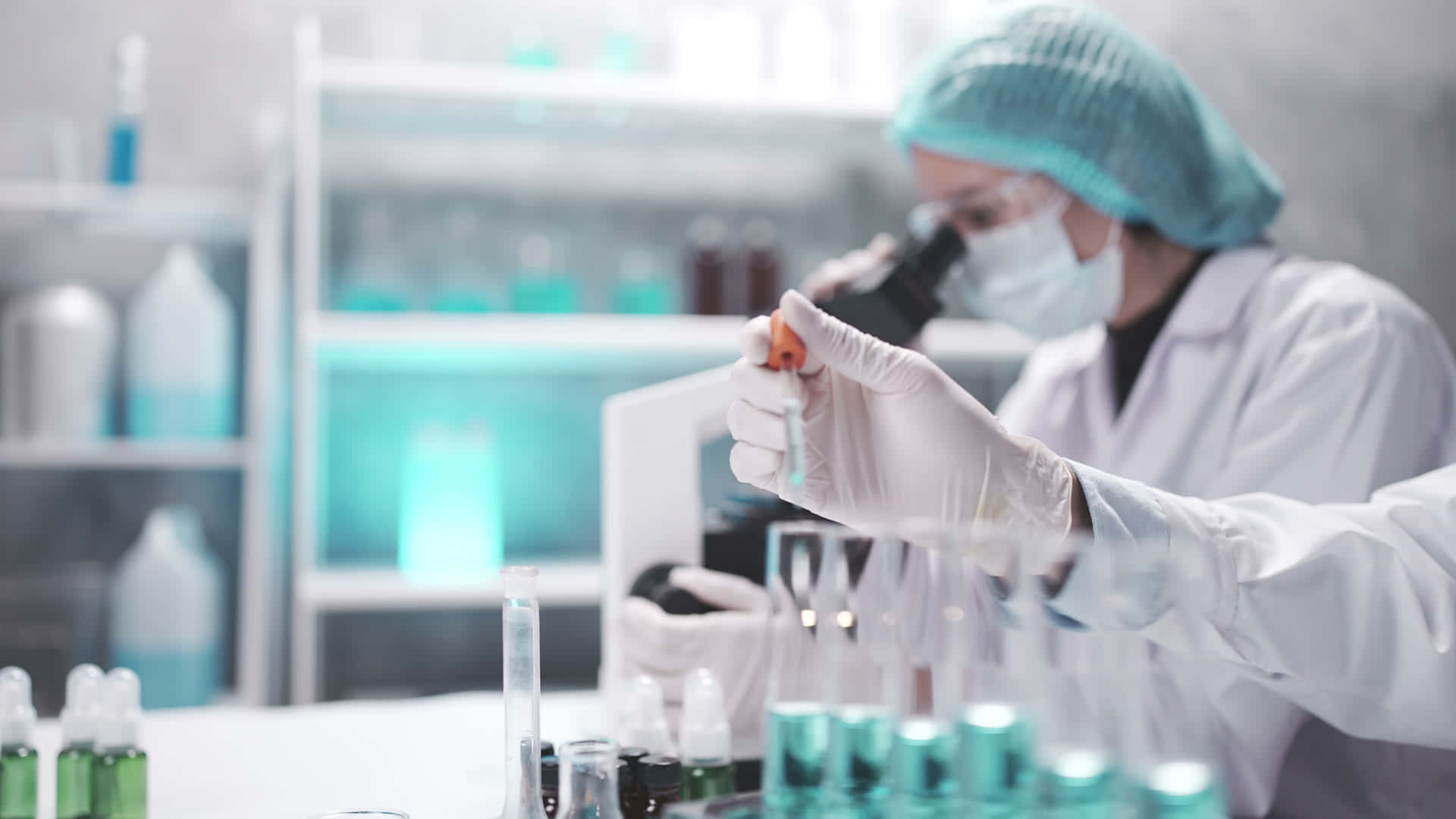 科學 桌布 壁紙 — 一位穿著實驗袍和口罩的科學家，在試管和瓶子環繞下，仔細地用顯微鏡檢查樣本。