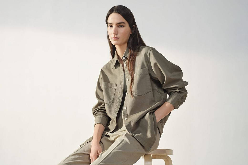 Uniqlo 桌布 壁紙 — 一位穿著寬鬆橄欖色襯衫和長褲的女性，自信地坐在白色背景前的木凳上。