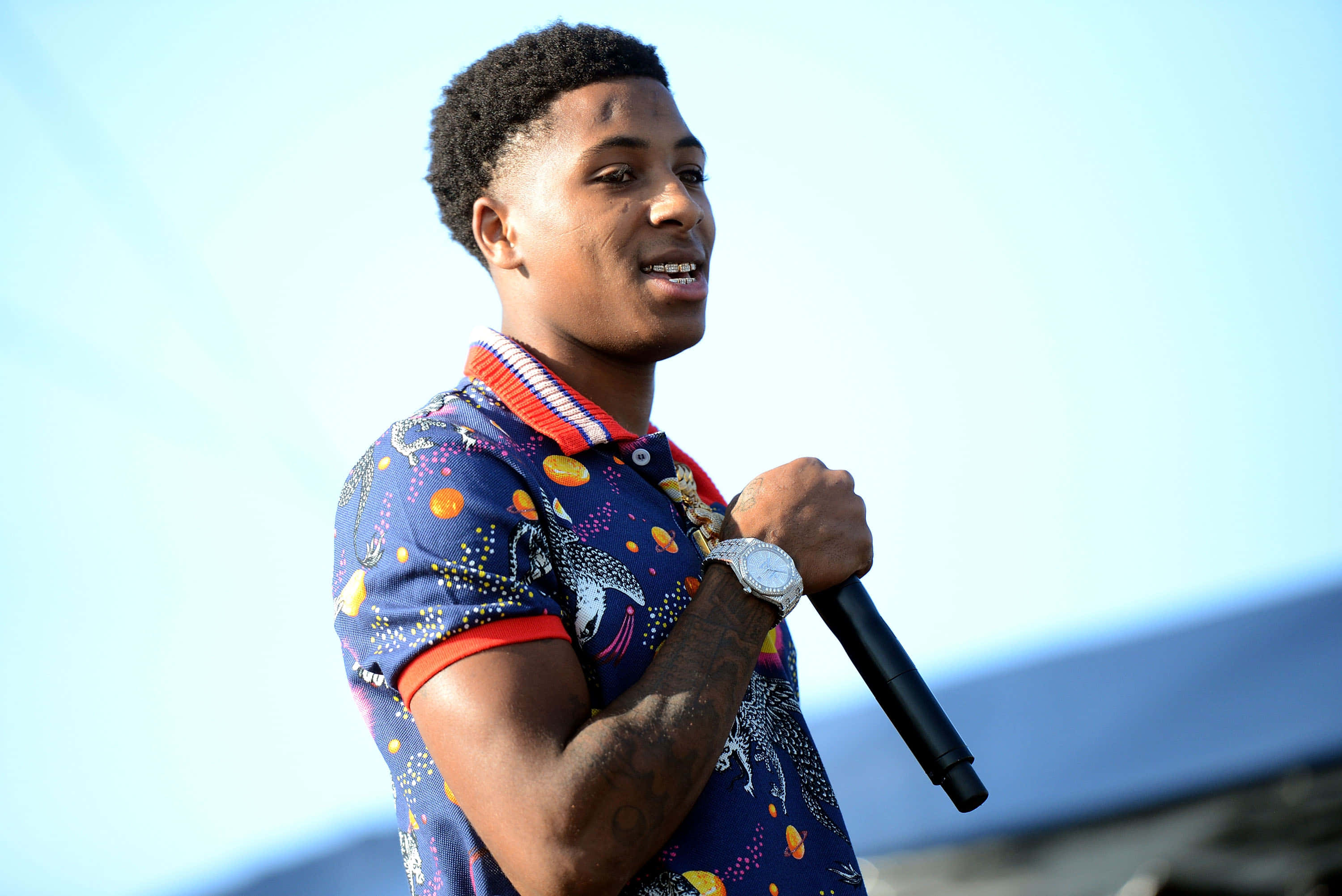 Youngboy 桌布 壁紙 — 一位穿著彩色圖案襯衫的年輕男子在明亮的藍天下拿著麥克風進行戶外表演。