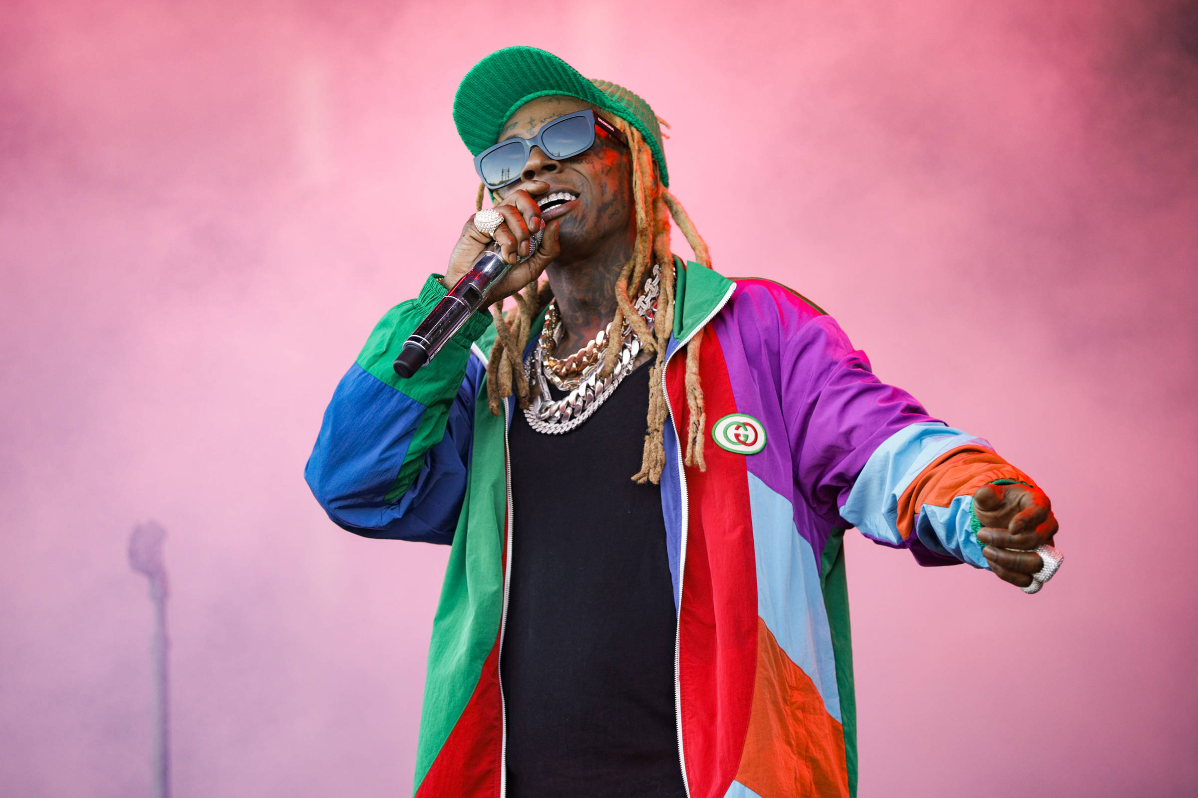Lil Wayne 桌布 壁紙 — 一位穿著彩色外套和綠色毛帽的饒舌歌手，在模糊的粉紅色煙霧背景下對著麥克風演唱。