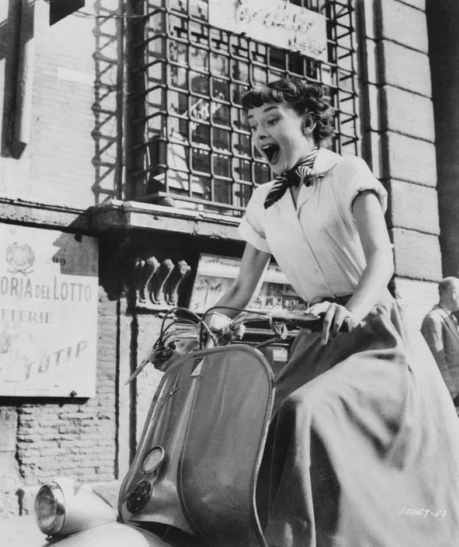Classic Movie 桌布 壁紙 — 一位穿著復古服裝的女性，滿懷喜悅地騎著滑板車行駛在陽光明媚的歐洲街道上。