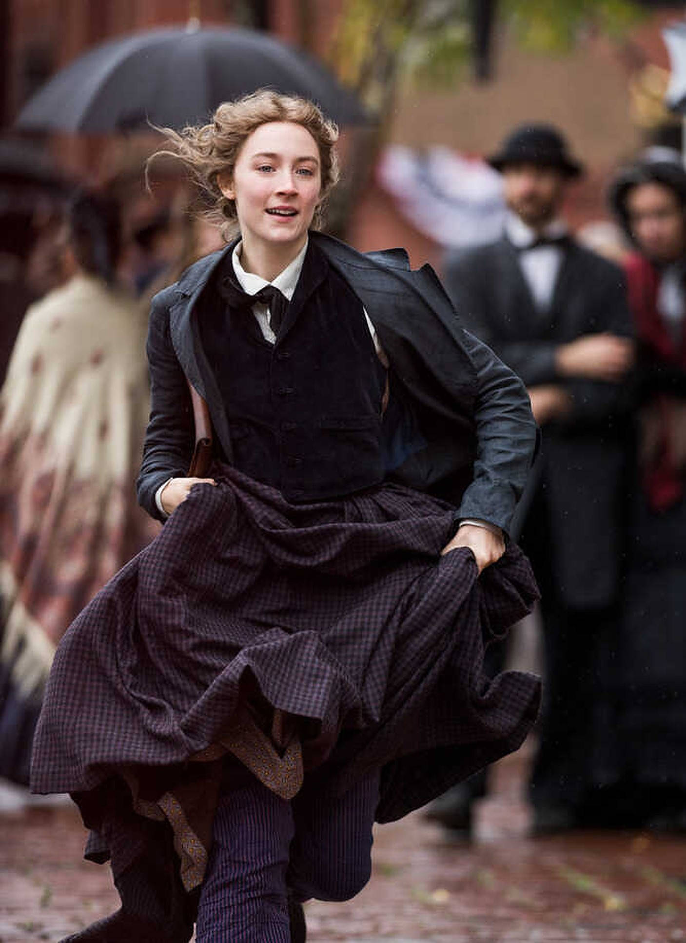 Saoirse Ronan 桌布 壁紙 — 一位穿著復古服裝的女性在雨中的鵝卵石街道上快樂地奔跑，旁觀者在傘下觀看。