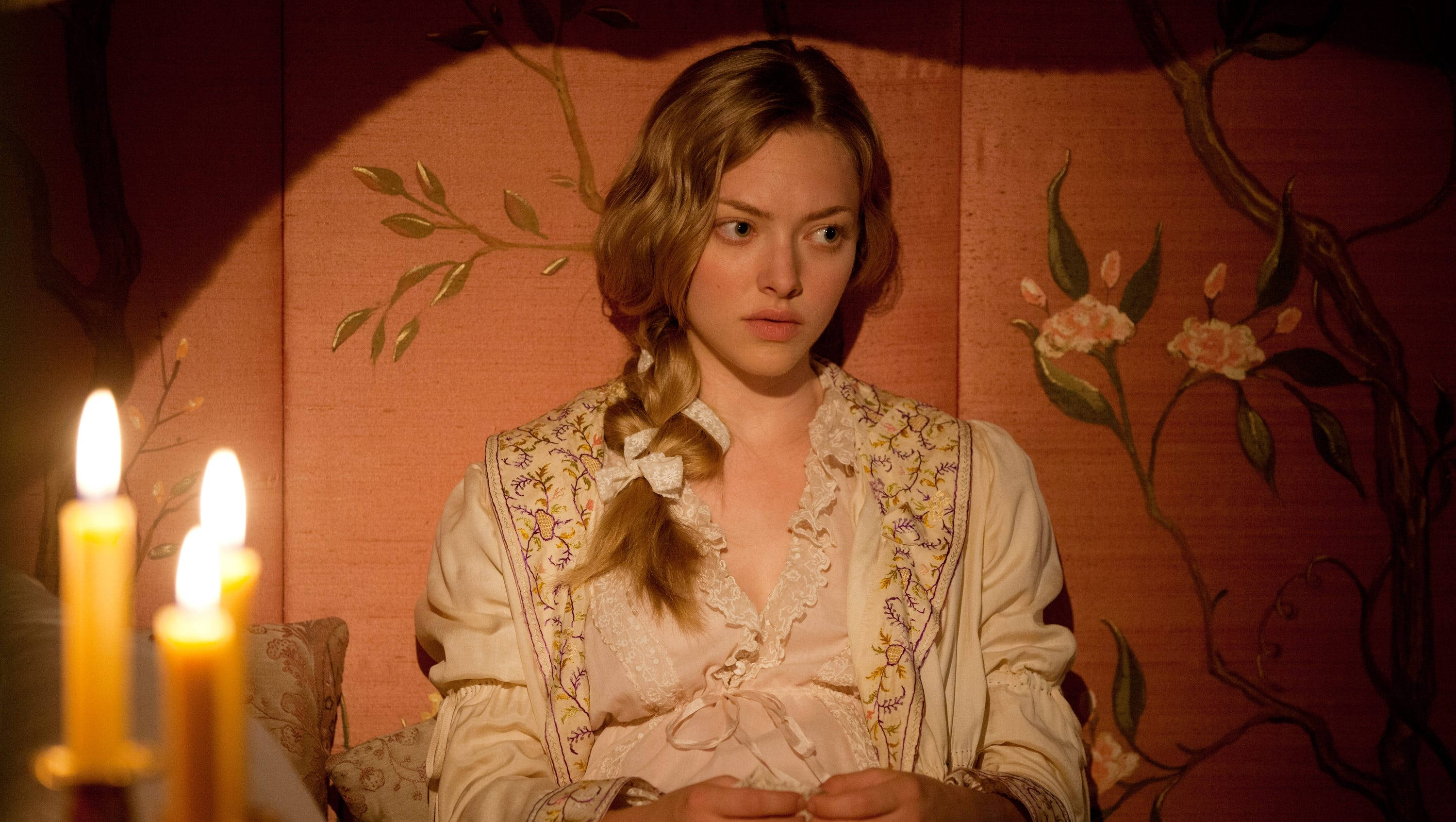 Amanda Seyfried 桌布 壁紙 — 一位穿著復古蕾絲服裝的年輕女性坐在一個溫暖照明的房間裡，燭光在花卉壁紙上投下柔和的陰影。