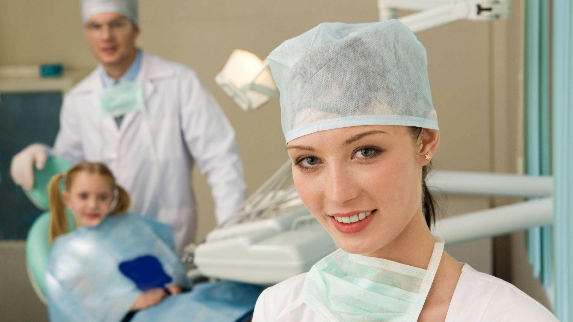 Dentistry 桌布 壁紙 — 一位穿著手術衣和帽子的微笑女牙醫看著鏡頭，而一個小女孩坐在牙科椅上，身後有另一位牙醫。