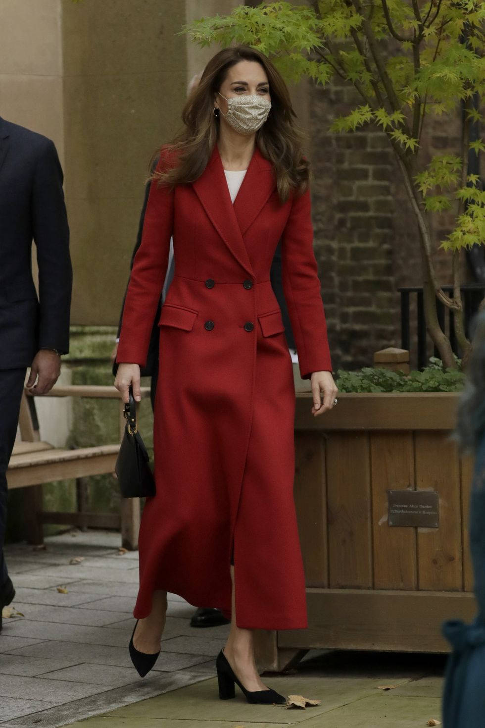 Kate Middleton 桌布 壁紙 — 一位穿著搶眼紅色雙排扣外套的女性在戶外行走，戴著有圖案的口罩和黑色高跟鞋。