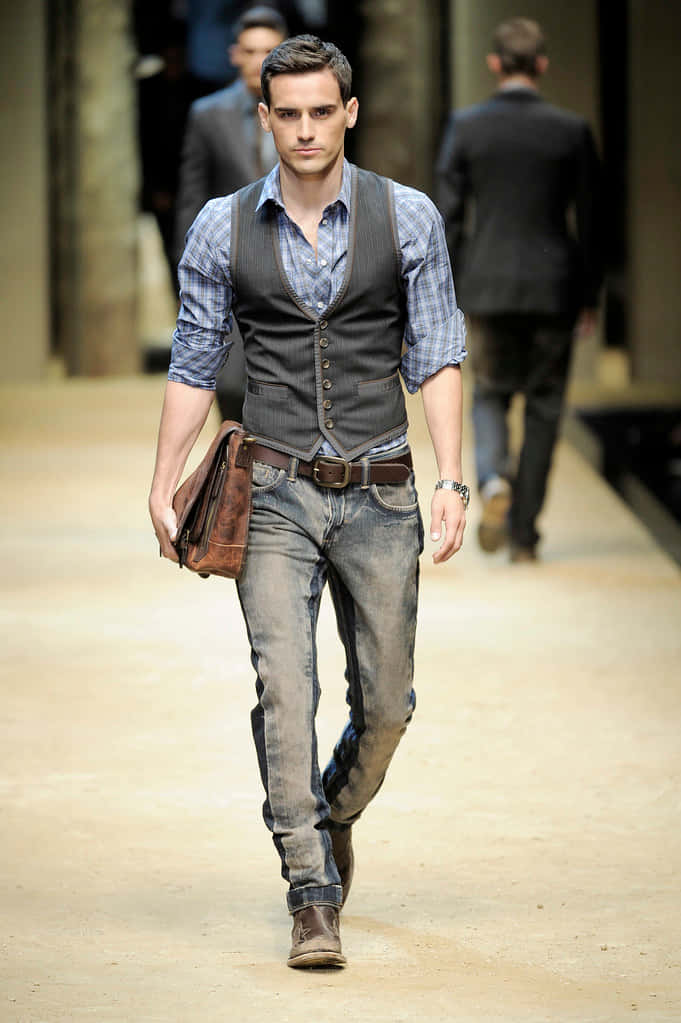 Mens Fashion 桌布 壁紙 — 一位穿著整齊的男士，身穿格紋襯衫、背心、牛仔褲和靴子，自信地走在時裝伸展台上，身後有模糊的模特兒。