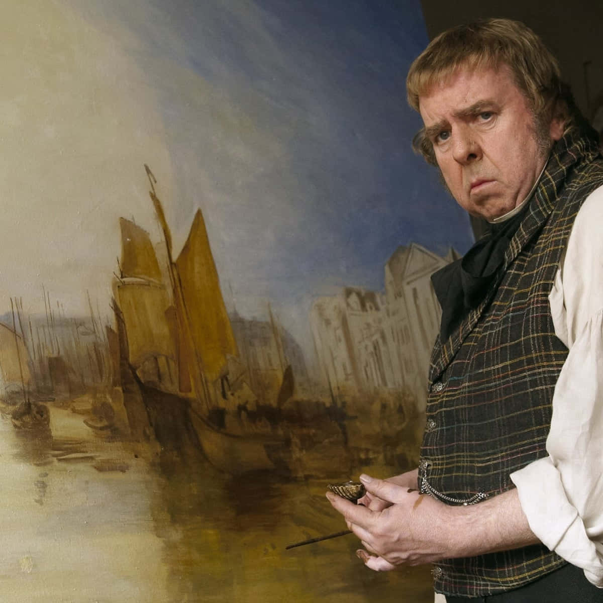 Timothy Spall 桌布 壁紙 — 一位穿著時代服裝的畫家手持畫筆，站在他描繪港灣船隻與建築的大型油畫旁。